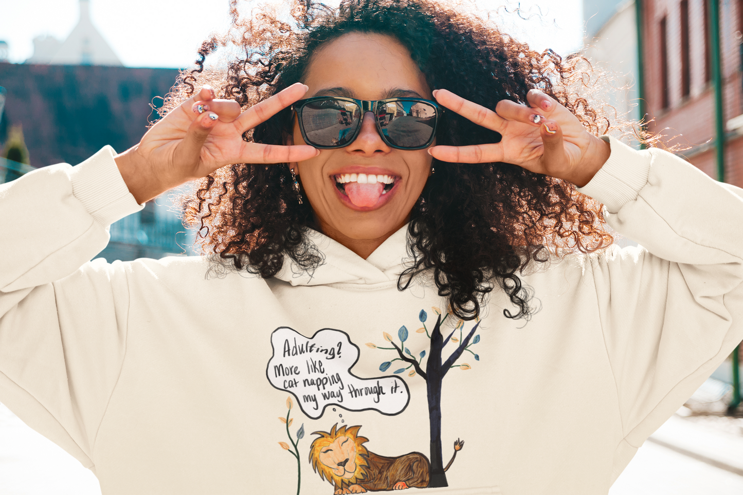Woman with curly hair and sunglasses sticking tongue out wearing oatmeal heather Adulting Nap Lion hoodie – sarcastic affirmation lazy vibes by GrungePup Sarcastic Affirmations