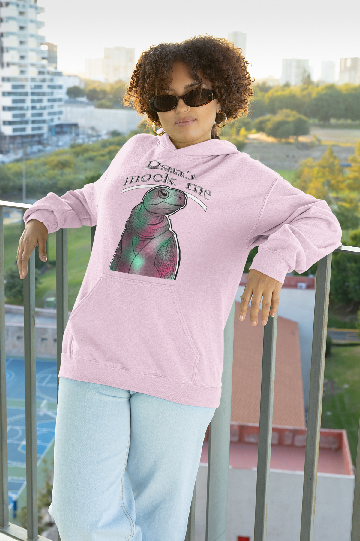 Woman with curly hair and sunglasses leaning on a balcony railing wearing a light pink Gildan hoodie with the Don’t Mock Me design