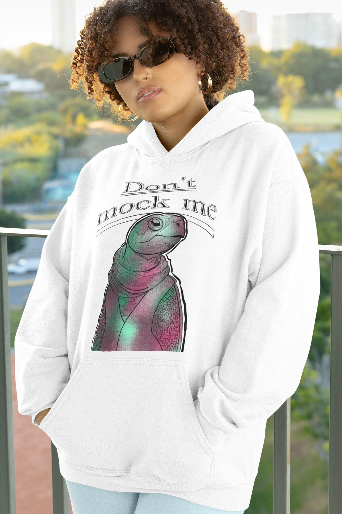 Woman with curly hair and sunglasses standing on a balcony wearing a white Gildan hoodie with the Don’t Mock Me Mock Turtle design