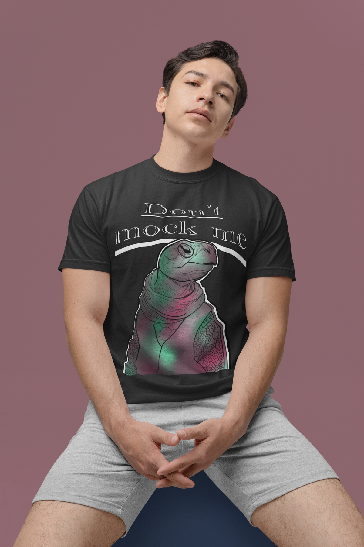 Serious young man sitting in a studio wearing a black Comfort Colors 1717 t-shirt with the Don’t Mock Me Mock Turtle design