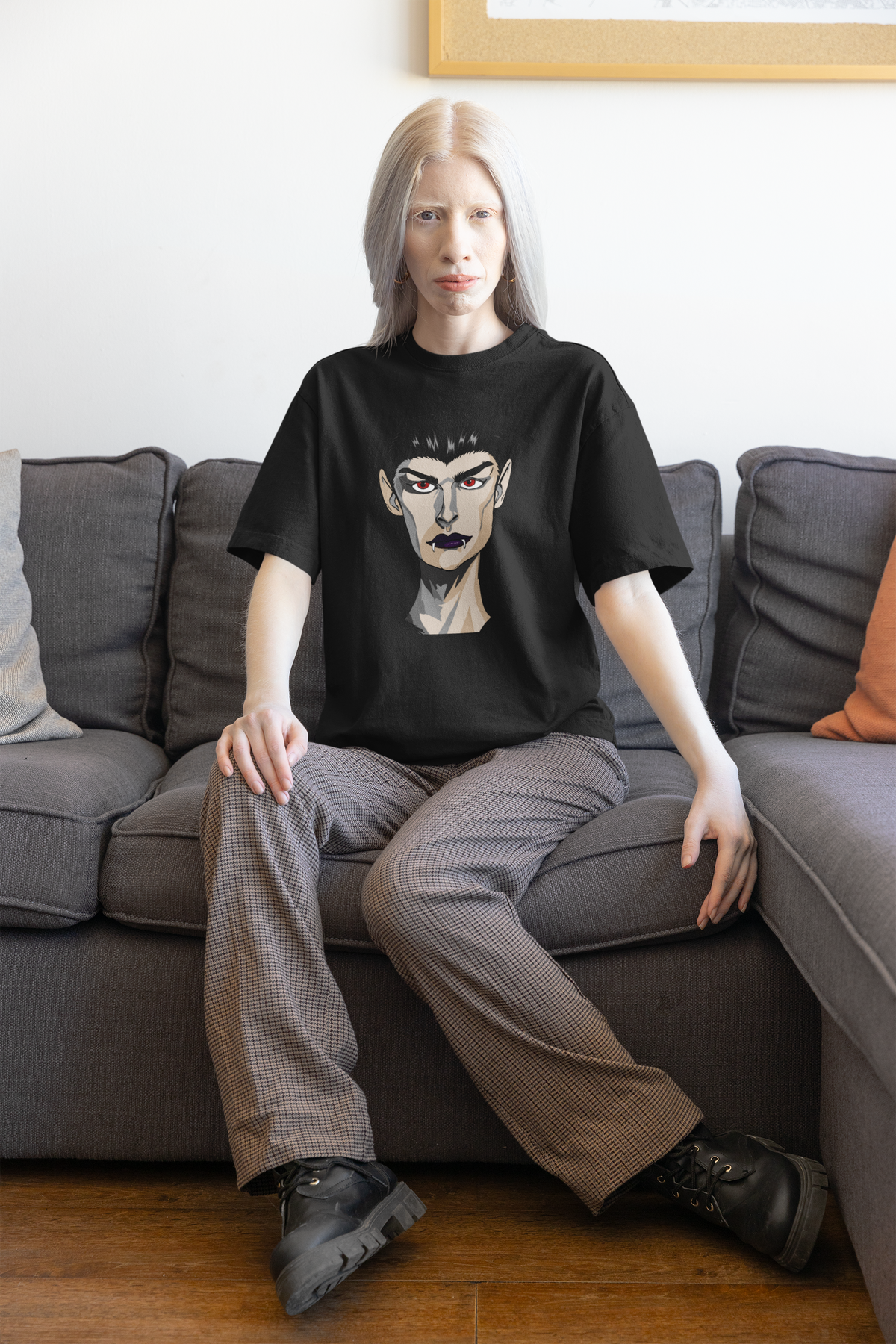 Woman with pale hair sitting on a gray sofa wearing a black heather t-shirt printed with the Emo Vamp vampire face.