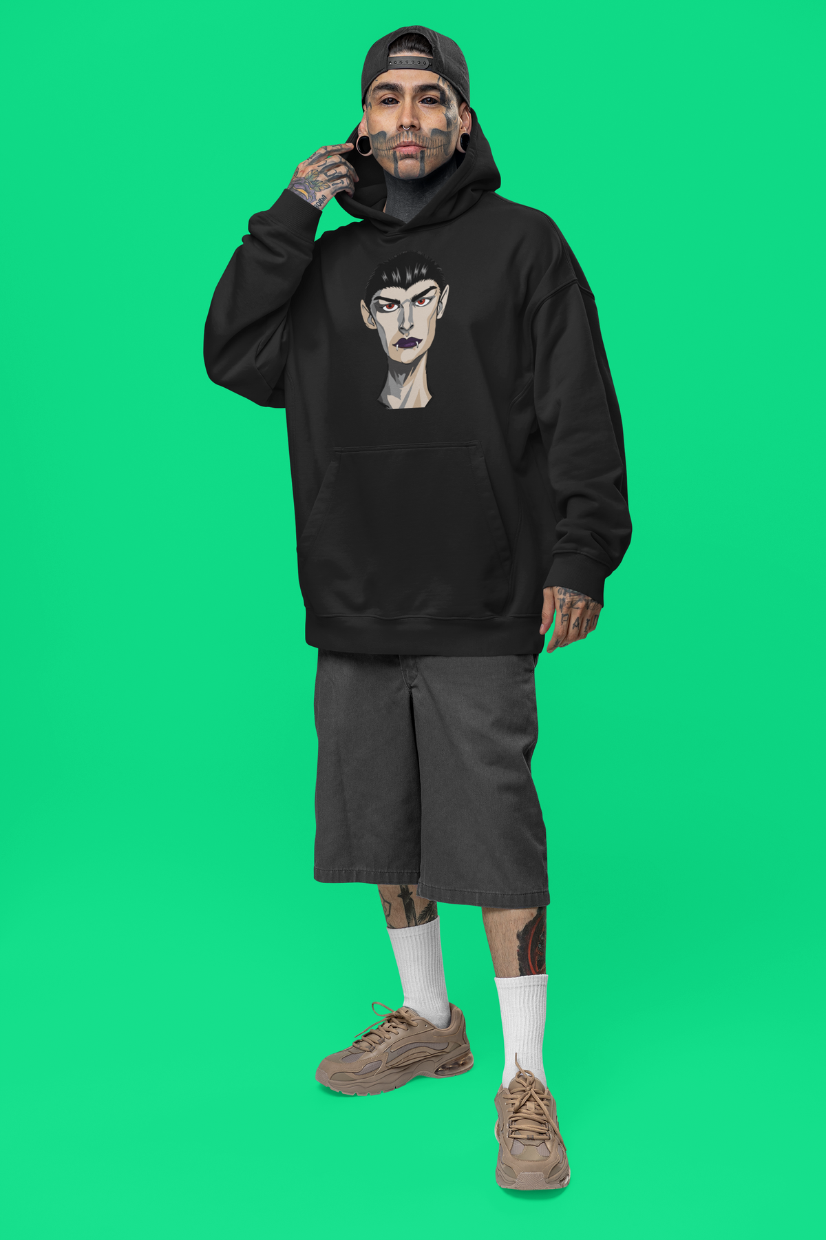 Tattooed man in a black cap wearing a black hoodie printed with the Emo Vamp vampire face against a green studio background.