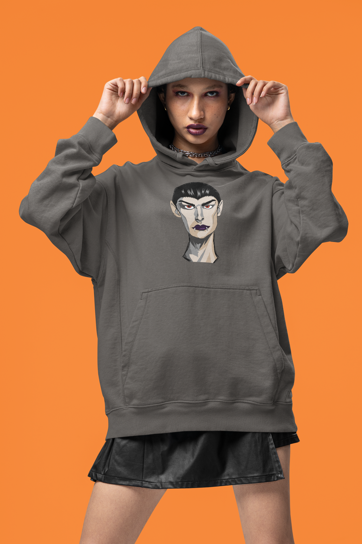 Woman pulling up the hood of a charcoal hoodie printed with the Emo Vamp vampire face against an orange background.
