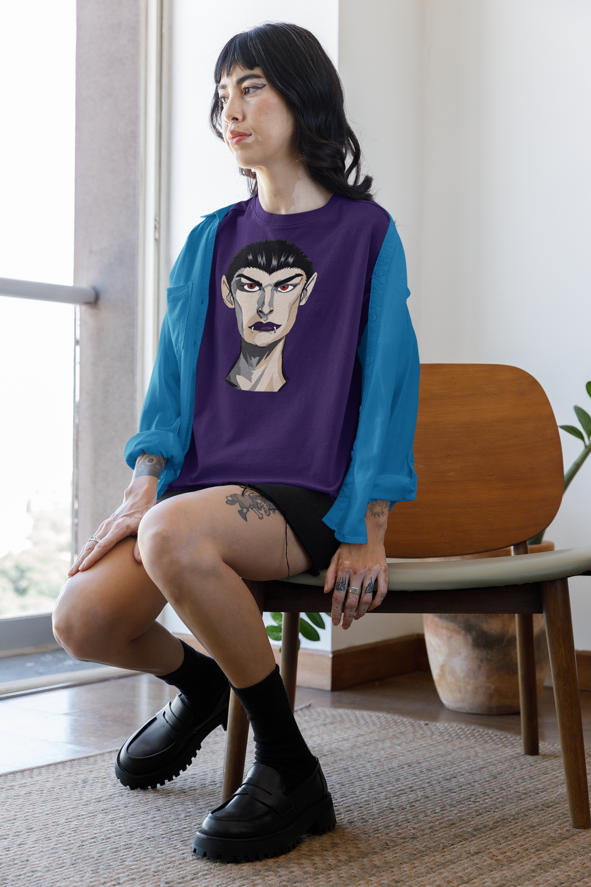 Tattooed woman sitting in a chair wearing a team purple t-shirt and an open blue shirt, printed with the Emo Vamp vampire face.