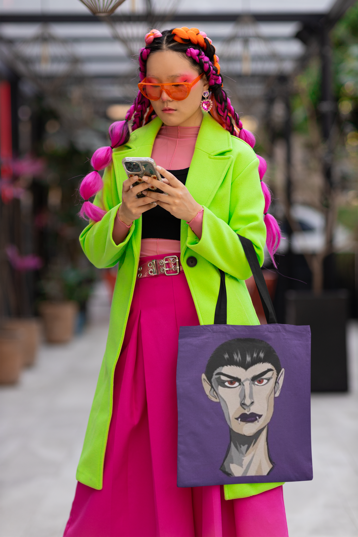 Woman with pink and orange braids and orange sunglasses carrying a purple tote bag printed with the Emo Vamp vampire face.