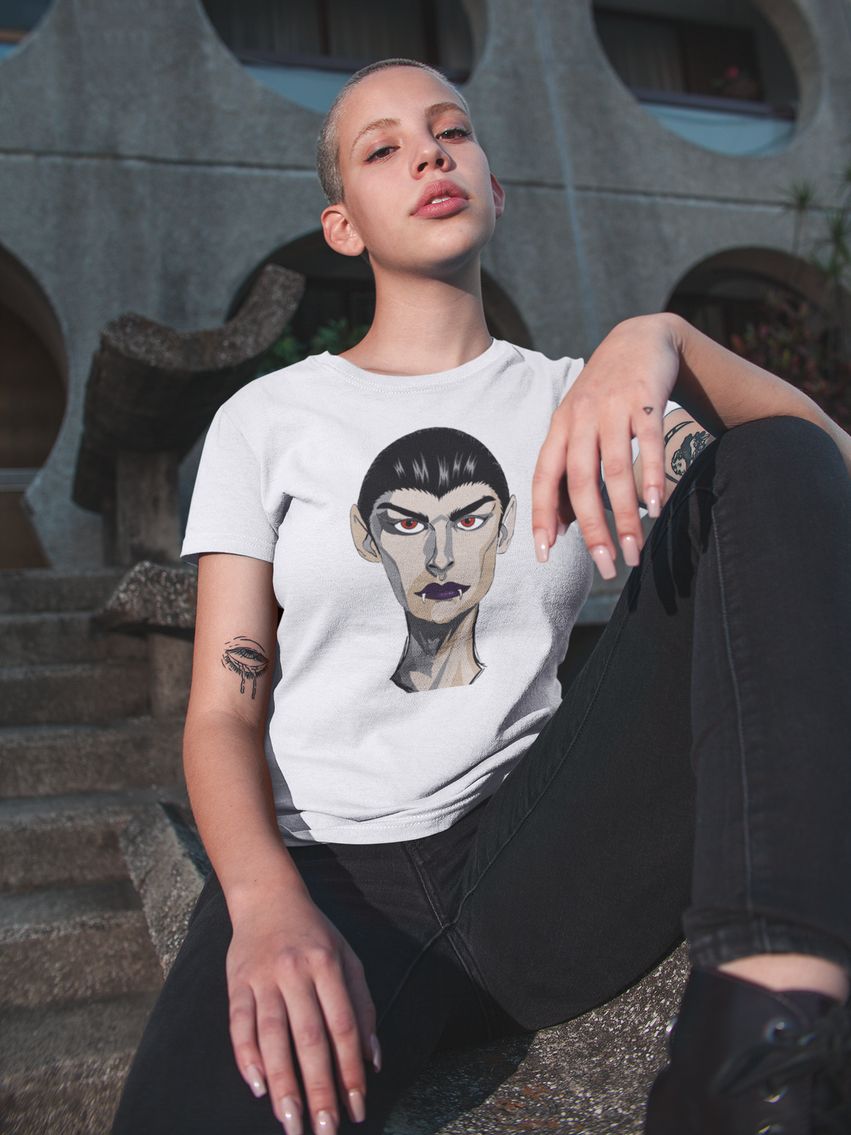 Woman with a buzz cut sitting on outdoor steps wearing a white t-shirt printed with the Emo Vamp vampire face.