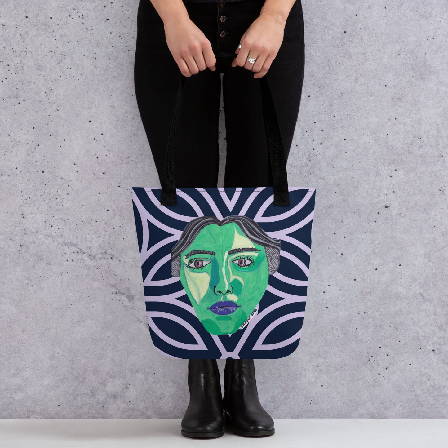 Person holding Envious Gaze tote bag with striking green face artwork on dark blue geometric pattern