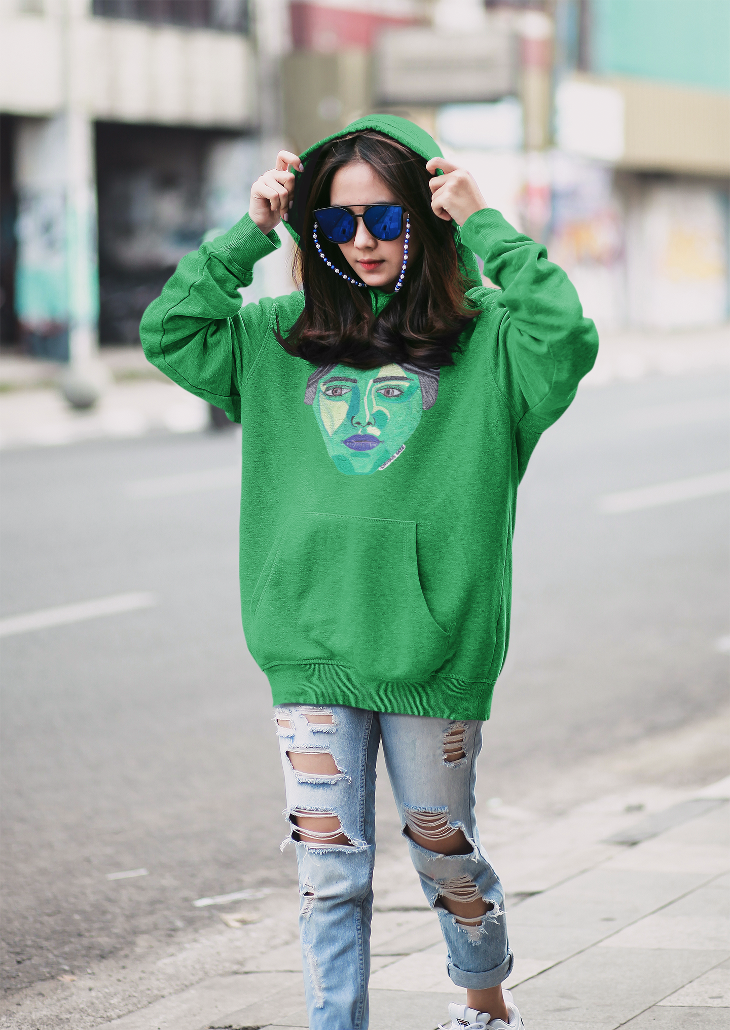 Woman wearing green Envious Gaze hoodie with colorful face design and ripped jeans standing on the street
