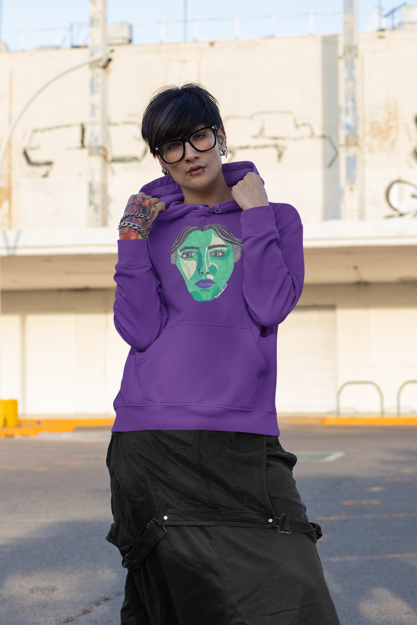 Androgynous woman wearing purple Envious Gaze hoodie with green face design in urban setting
