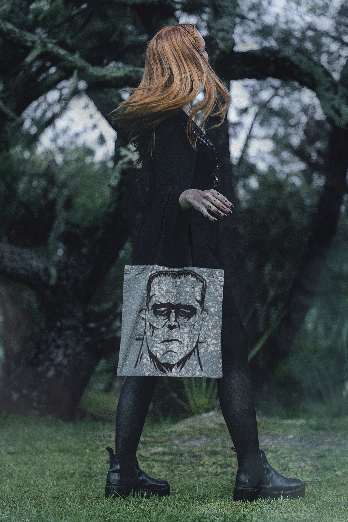 Woman with long red hair wearing black outfit carrying Frank's Monster all-over print tote bag in a forest setting