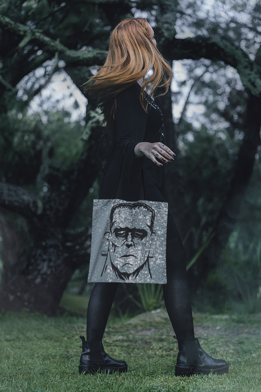 Woman with long red hair wearing black outfit carrying Frank's Monster all-over print tote bag in a forest setting