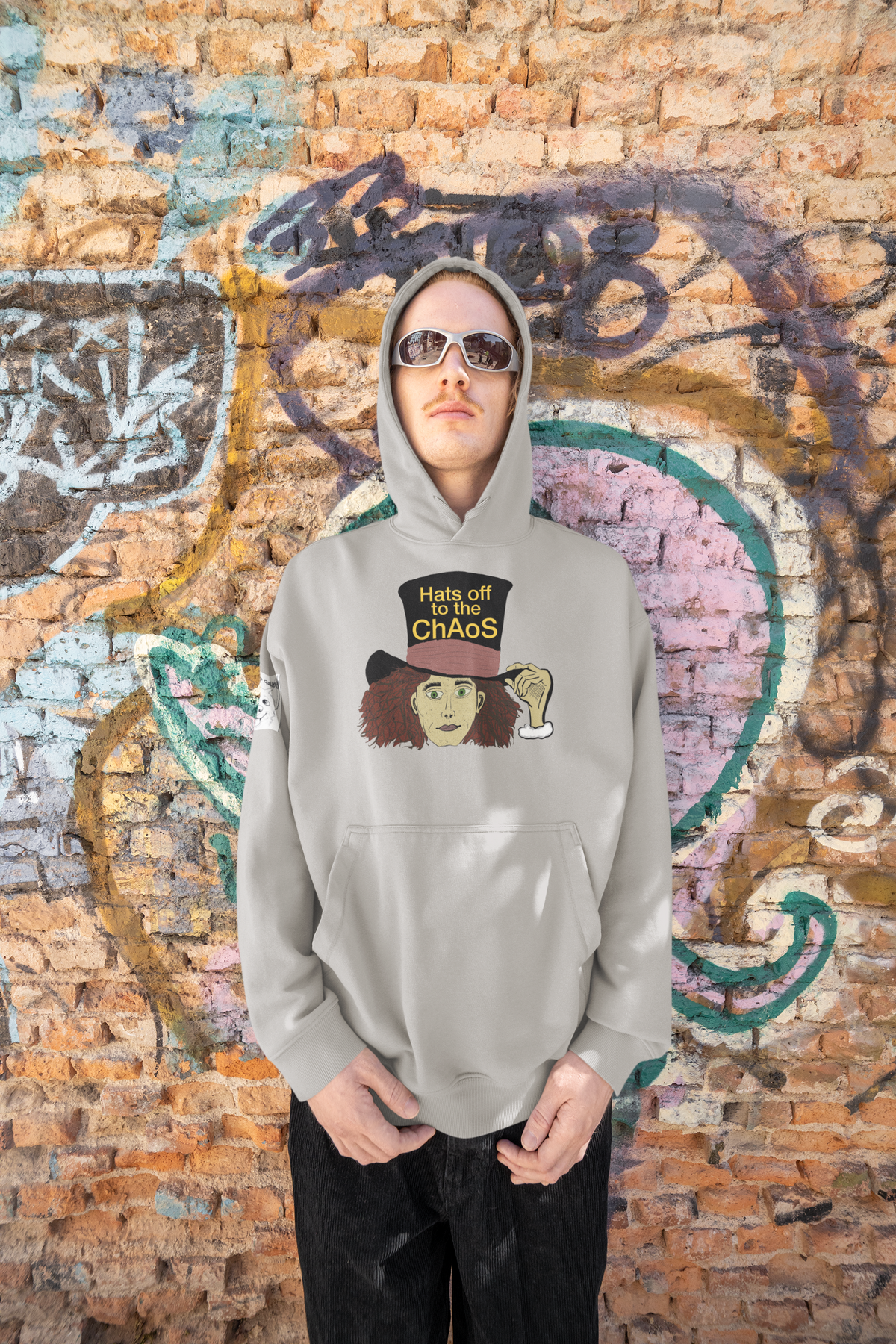 Man in sunglasses and a carbon grey Hats Off to the Chaos Mad Hatter hoodie in front of a brick graffiti wall. That Girl, Alice by GrungePup Gallery.