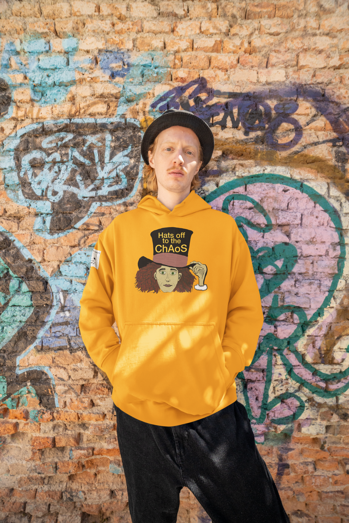 Man in a black hat and team gold Hats Off to the Chaos Mad Hatter hoodie in front of a brick graffiti wall. That Girl, Alice by GrungePup Gallery.