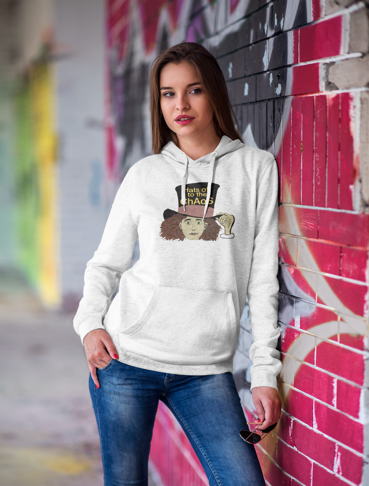 Woman standing against a pink brick wall in a white Hats Off to the Chaos Mad Hatter hoodie. That Girl, Alice by GrungePup Gallery.