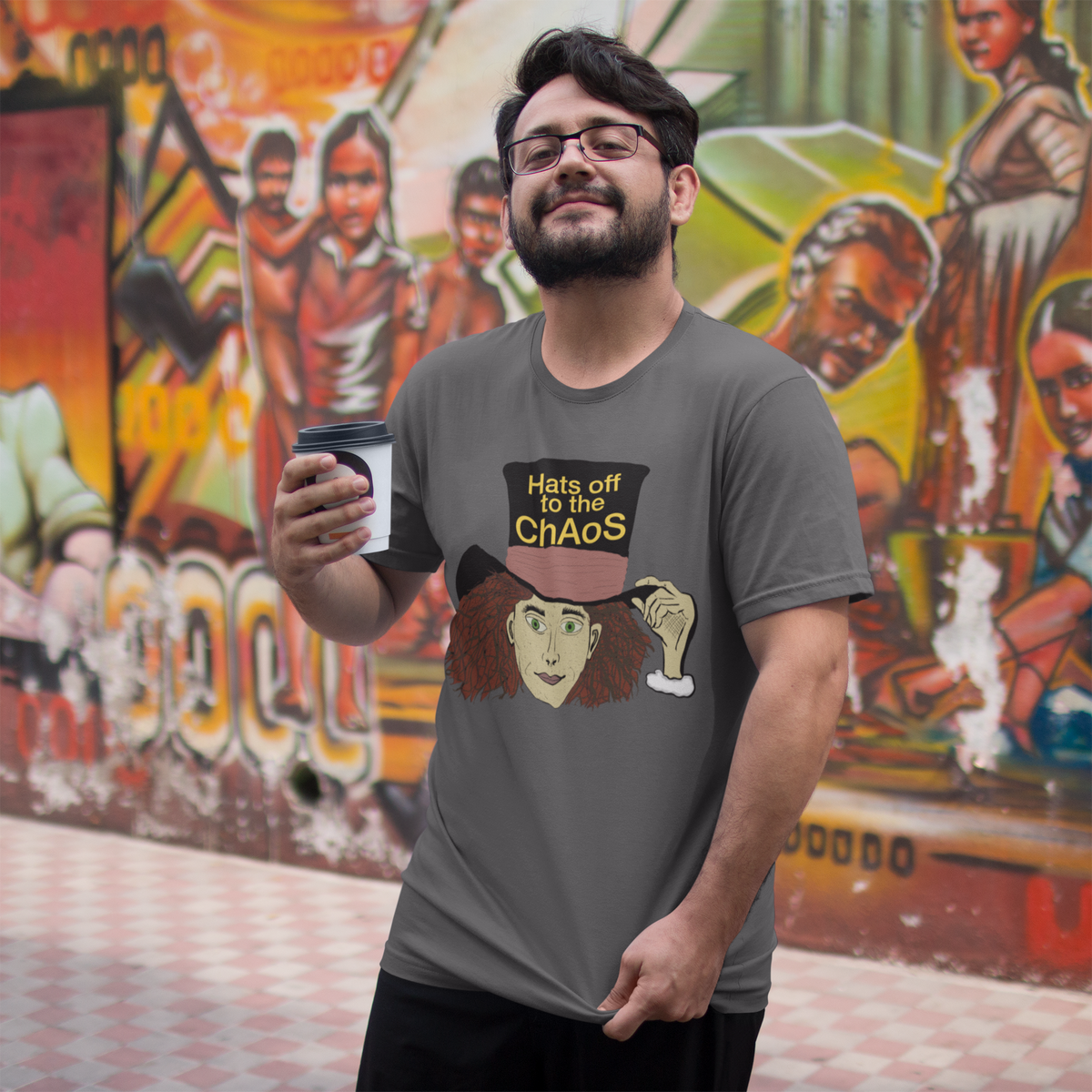 Bearded man in glasses holding coffee in a dark heather Hats Off to the Chaos Mad Hatter t-shirt in front of a mural. That Girl, Alice by GrungePup Gallery.