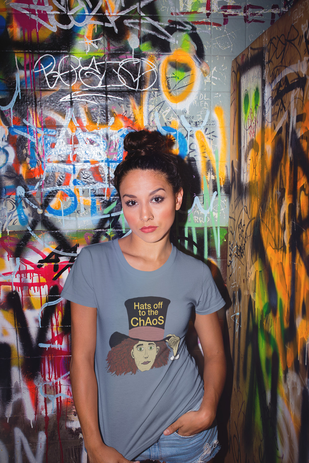 Woman with a bun in a heather indigo Hats Off to the Chaos Mad Hatter t-shirt standing in a graffiti-covered bathroom. That Girl, Alice by GrungePup Gallery.