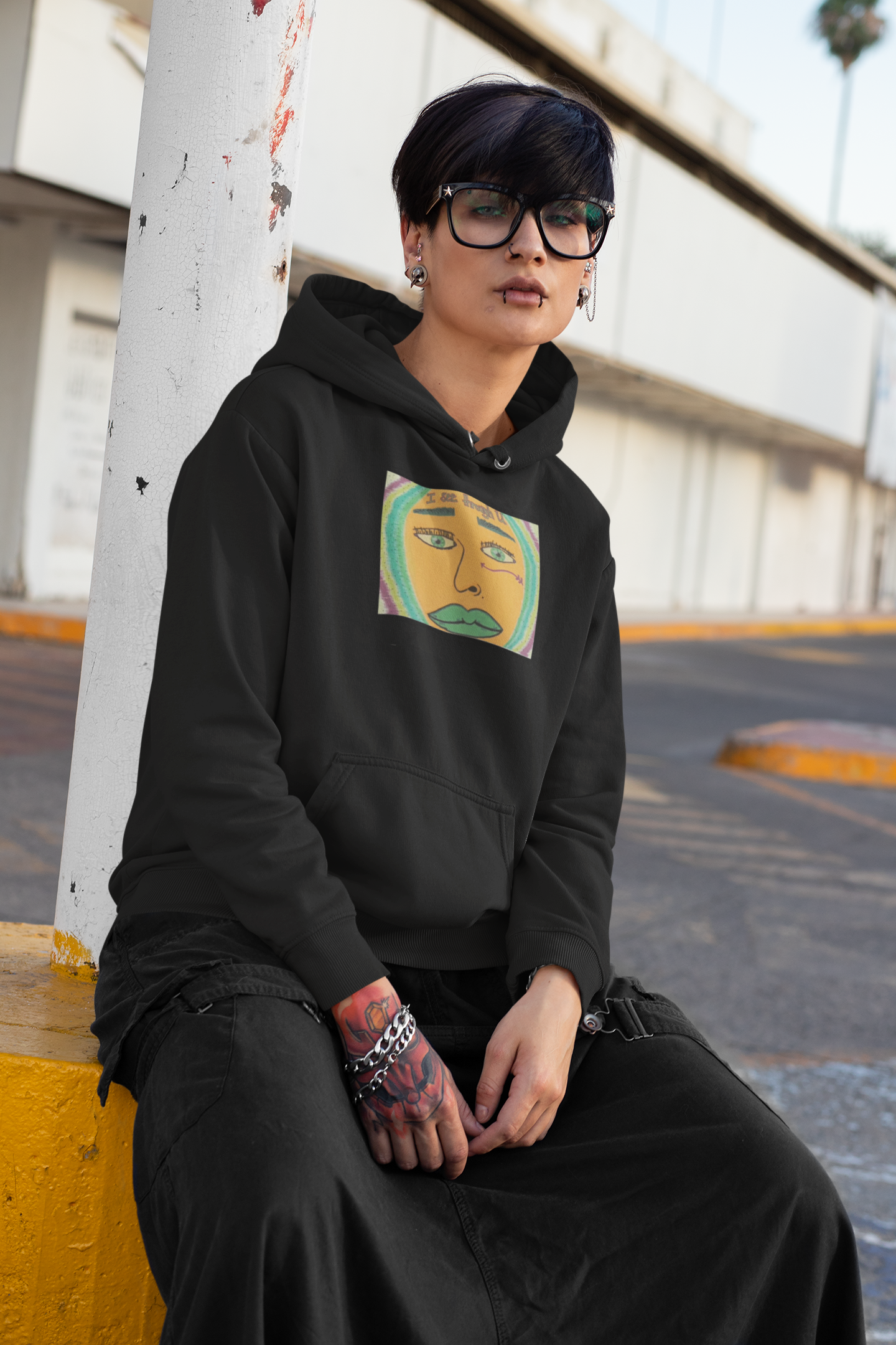 Androgynous person with short hair and piercings wearing black I See Through U hoodie leaning against pole
