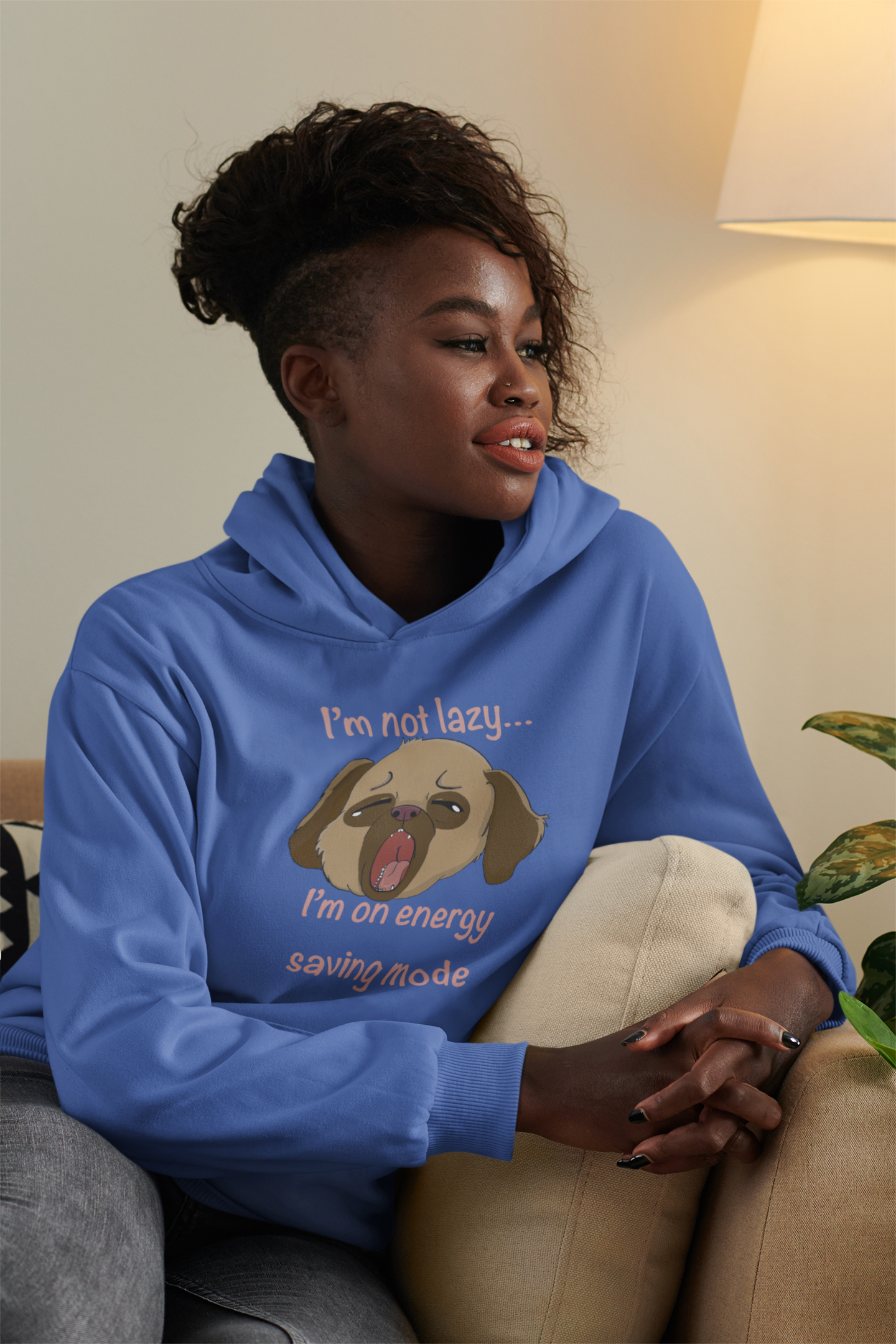 Woman with curly hair sitting on a couch wearing indigo blue I'm Not Lazy... I'm On Energy Saving Mode hoodie with sleepy pup design