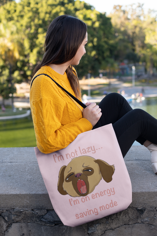 Woman in yellow sweater sitting outdoors by the water holding the pink I'm Not Lazy... I'm On Energy Saving Mode tote bag with tired pup design