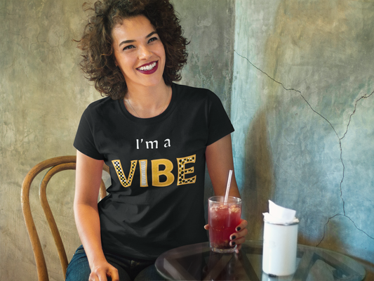 Smiling woman in the black heather I'm a Vibe tee holds a red drink at a bar.