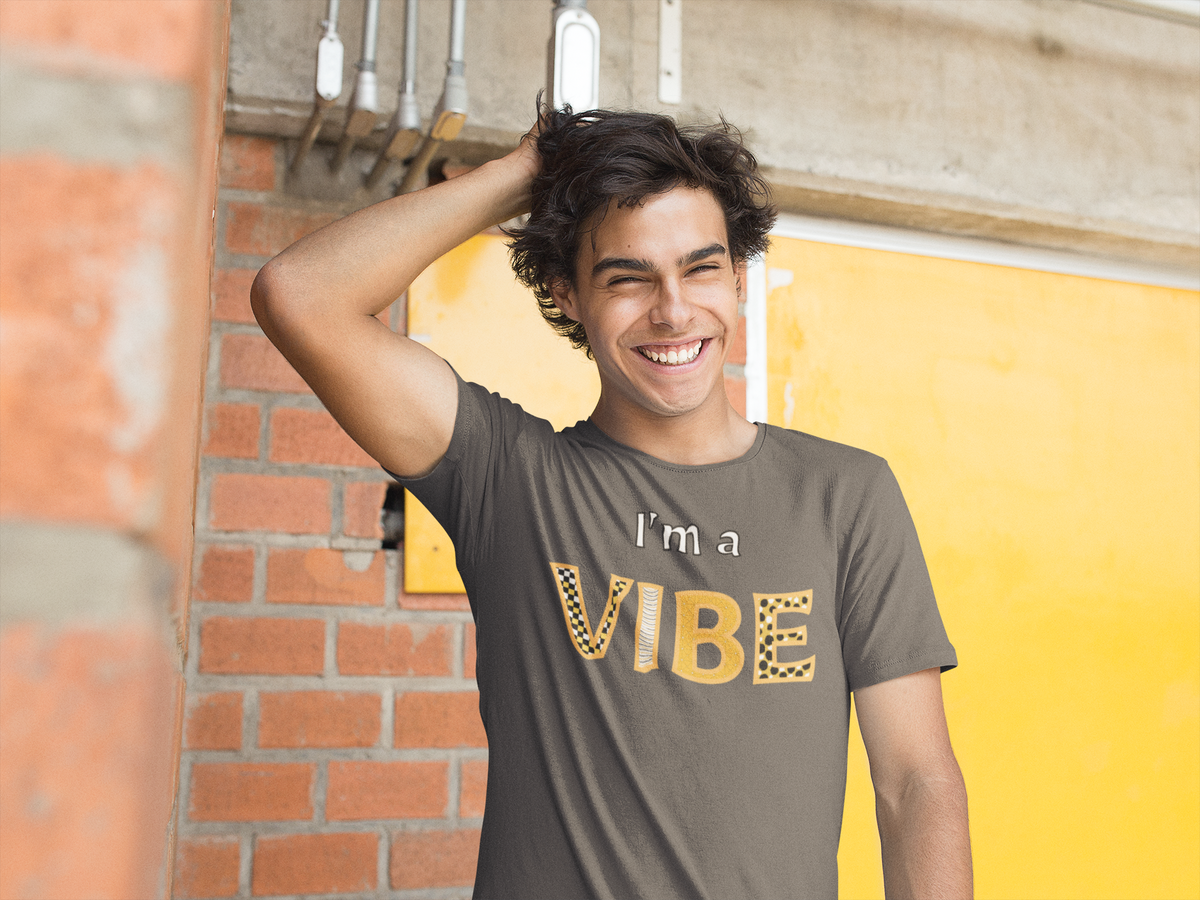 Smiling man with a hand in his hair wears the dark grey heather I'm a Vibe tee against a brick and yellow wall.
