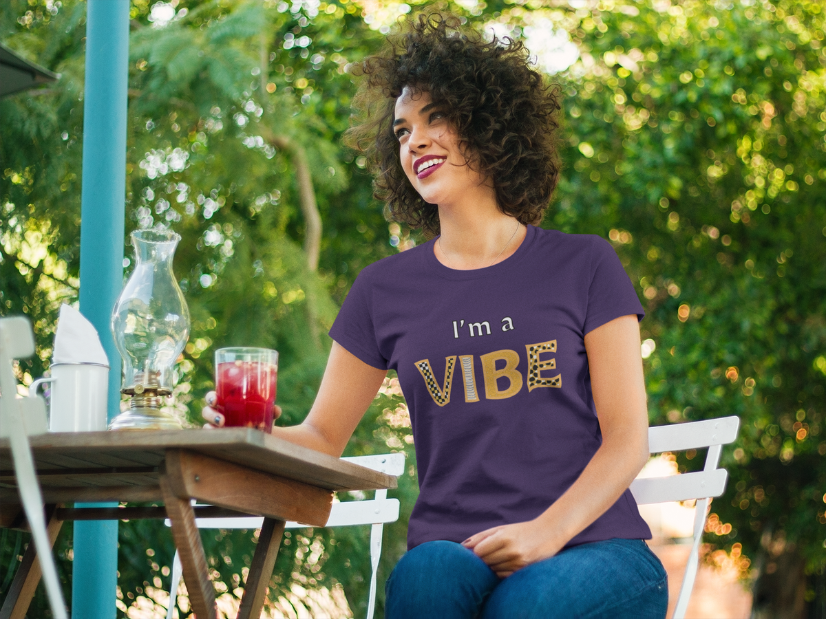 Smiling woman in the team purple I'm a Vibe tee holds a red drink at an outdoor table.