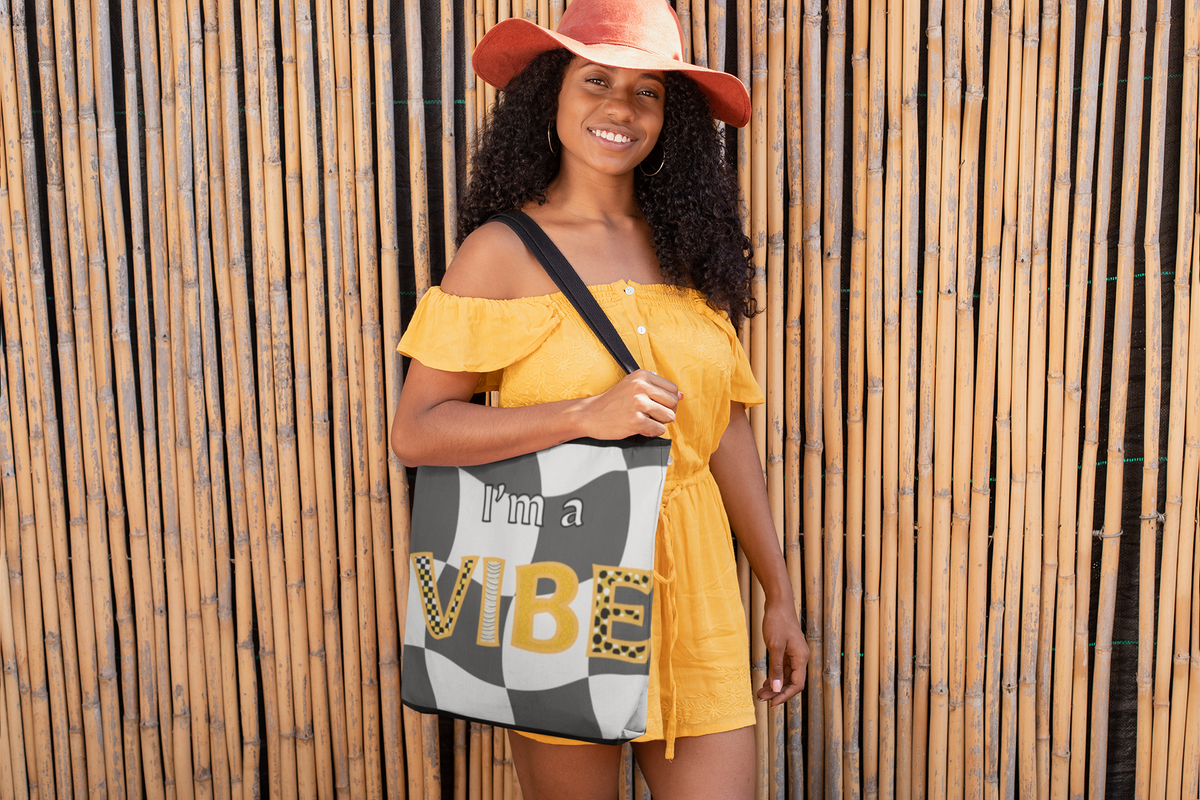 Smiling woman in a yellow off-shoulder dress and coral hat holds the grey checker I'm a Vibe tote against a bamboo fence.