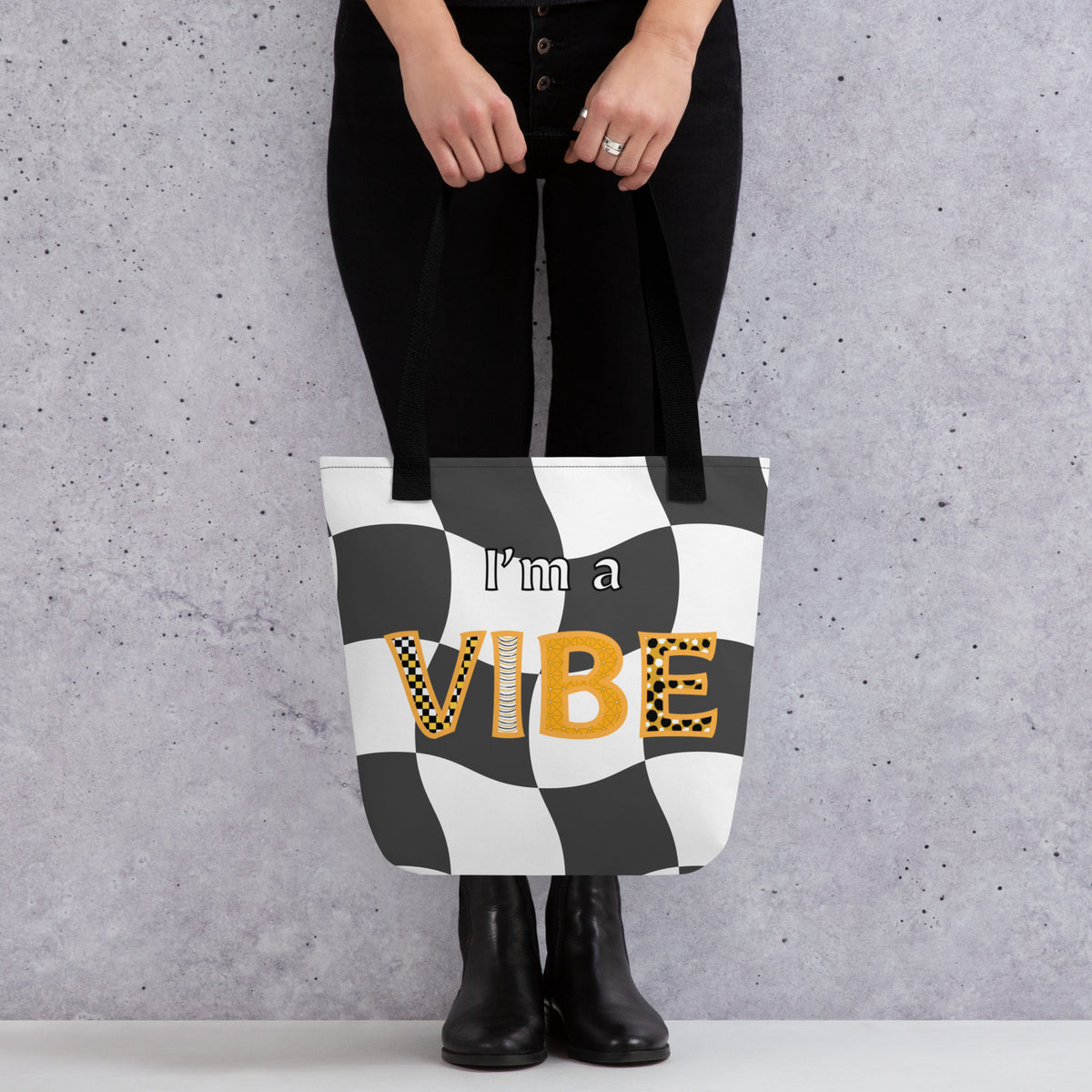 Woman from the waist down holds the front of the grey checker I'm a Vibe tote by its black straps.