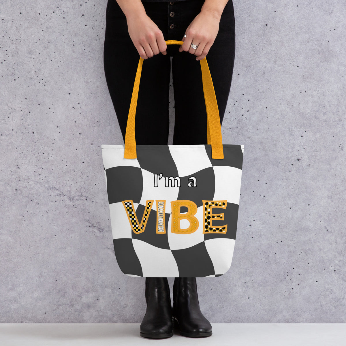 Woman from the waist down holds the front of the grey checker I'm a Vibe tote by its yellow straps.