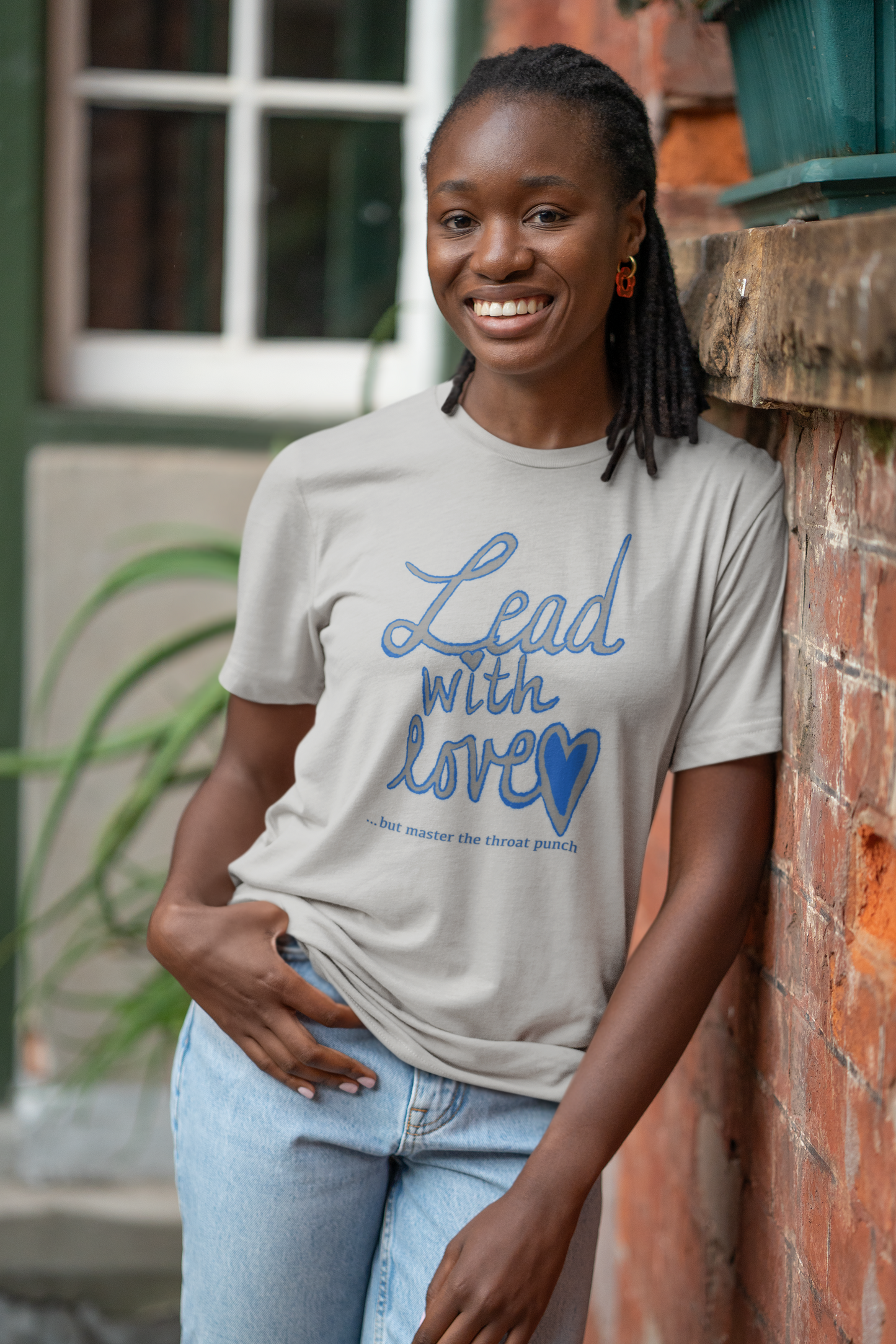 Smiling woman leaning against brick wall in athletic heather Bella Canvas t-shirt with "Lead with Love… but master the throat punch" sarcastic design. Bold self-care message for strong boundaries.
