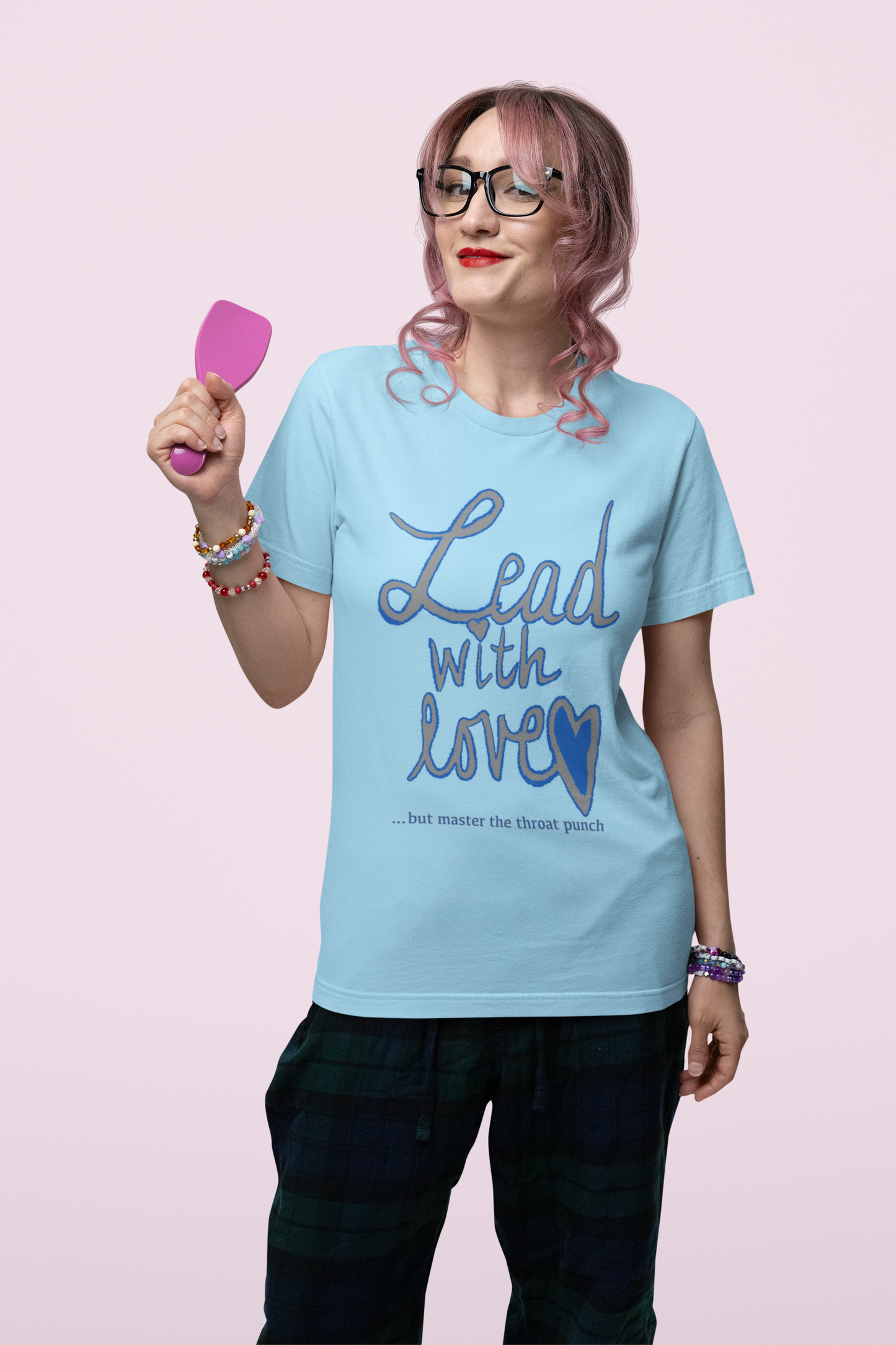 Woman with pink hair and glasses holding hairbrush in ocean blue Bella Canvas t-shirt with "Lead with Love… but master the throat punch" sarcastic design. Playful and empowering self-care message.