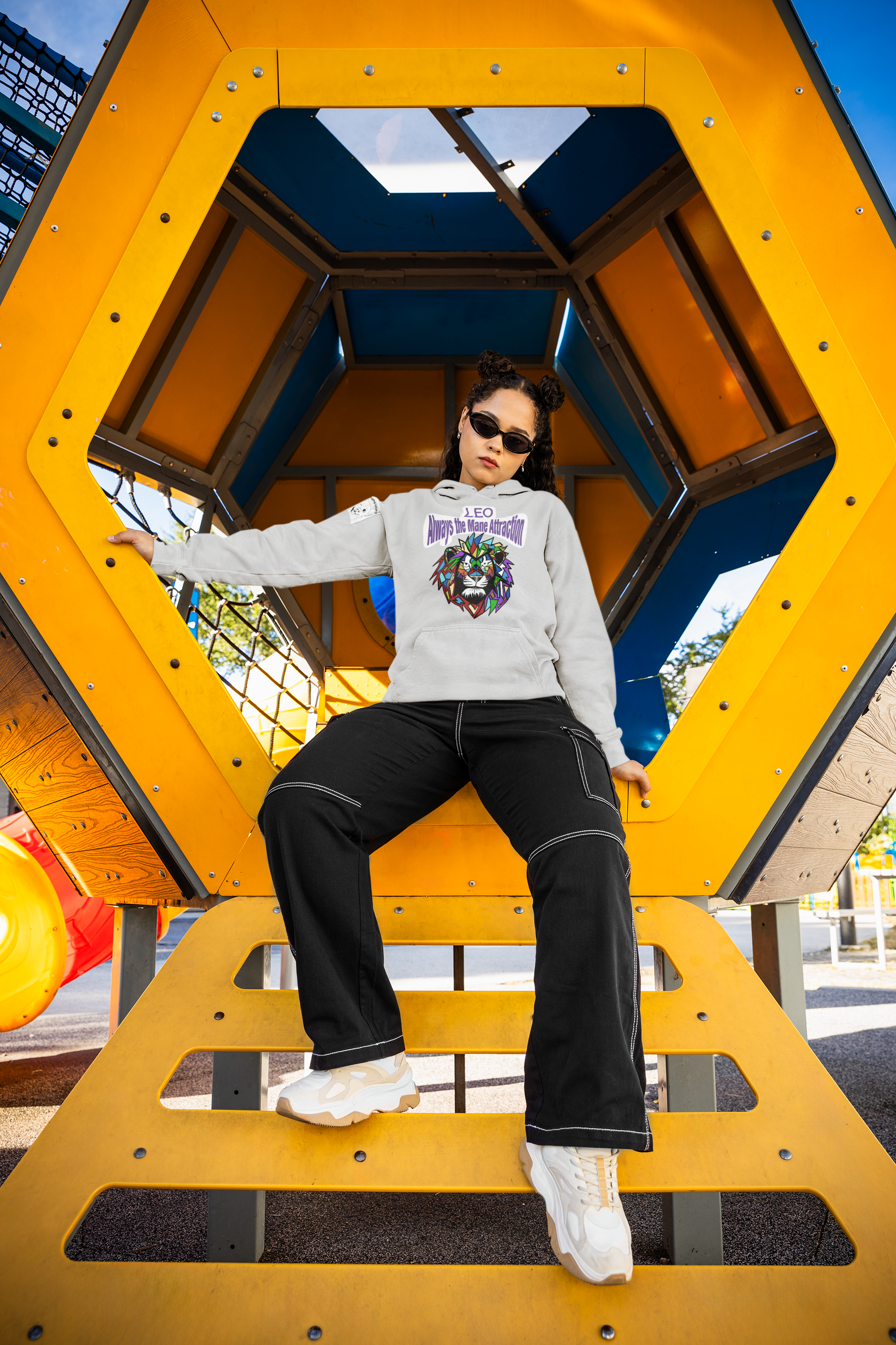 Woman with sunglasses and space buns posing inside a yellow playground structure while wearing an ash Leo Always the Mane Attraction hoodie