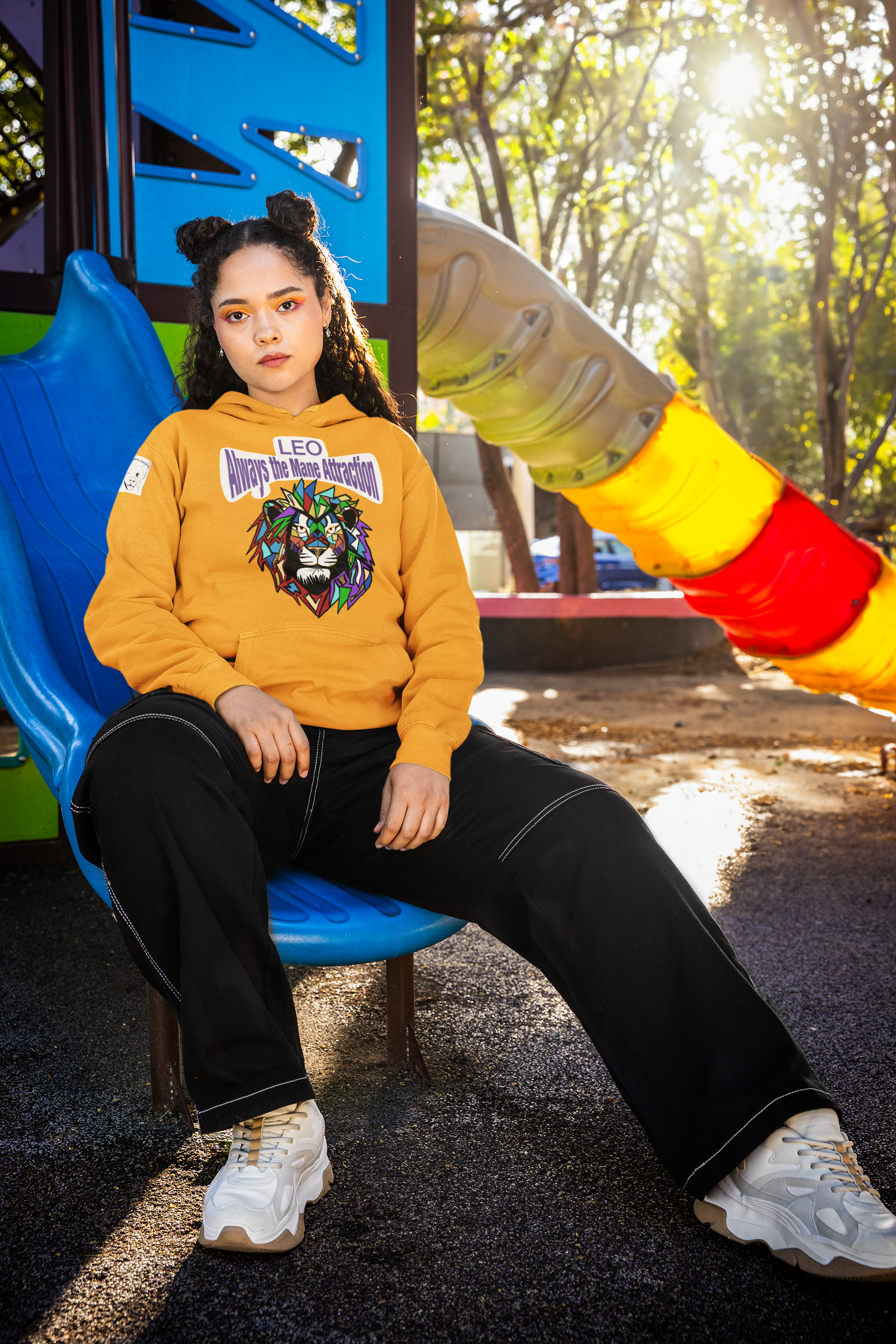 Woman with space buns sitting on a blue playground slide wearing a gold Leo Always the Mane Attraction hoodie in a sunny outdoor park
