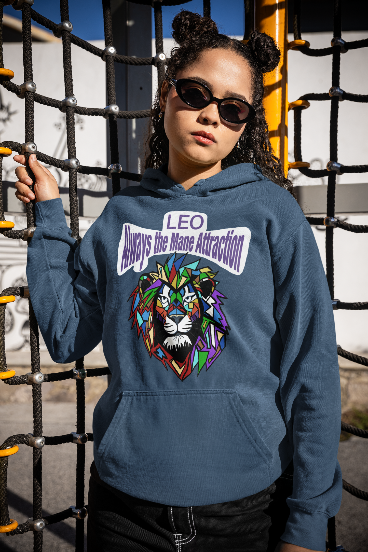 Woman with curly hair in space buns and sunglasses wearing an indigo blue Leo Always the Mane Attraction hoodie while standing by a playground climbing net