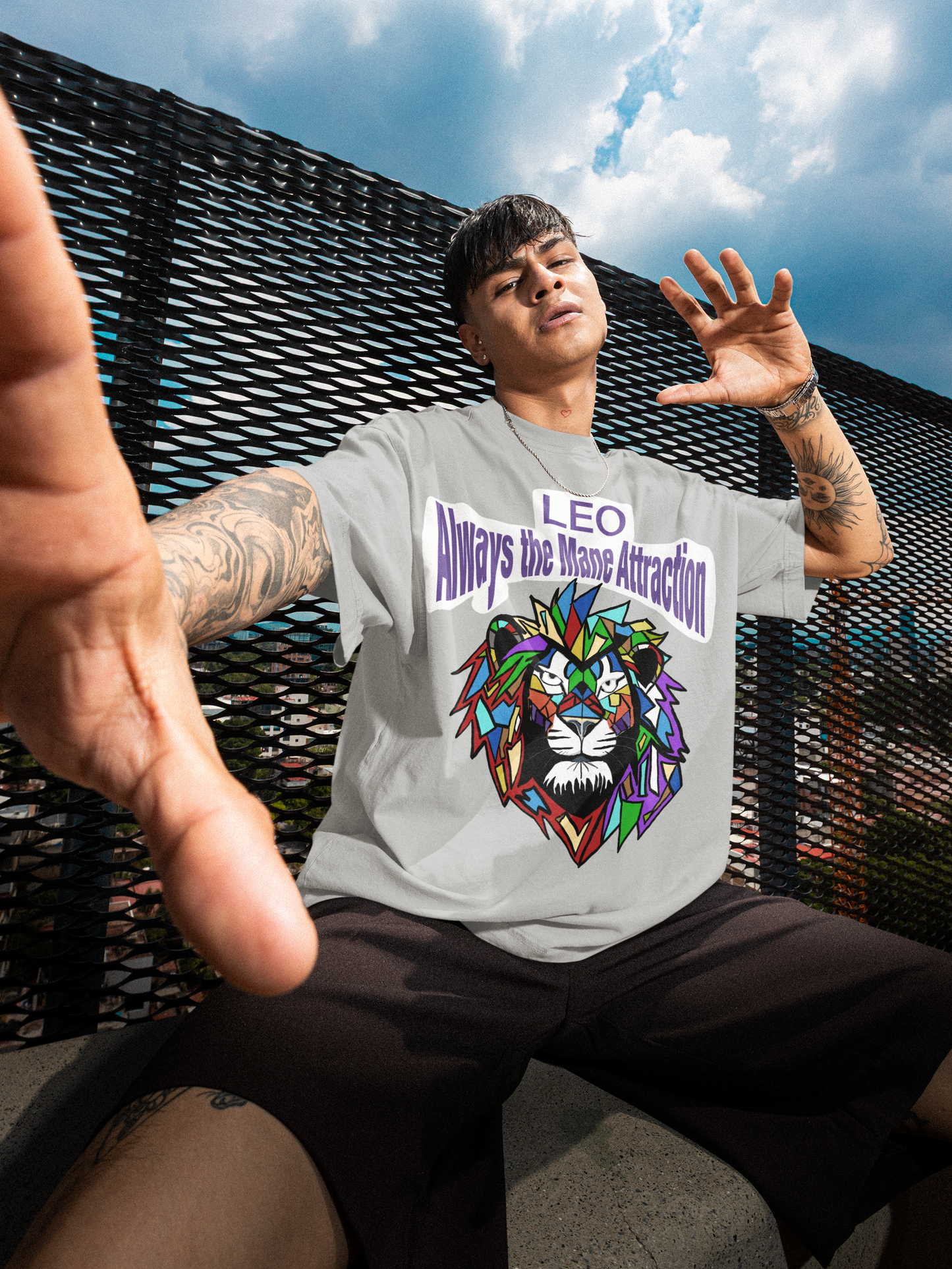 Tattooed man sitting on a rooftop with arms outstretched, wearing an athletic heather Leo Always the Mane Attraction t-shirt under a cloudy sky