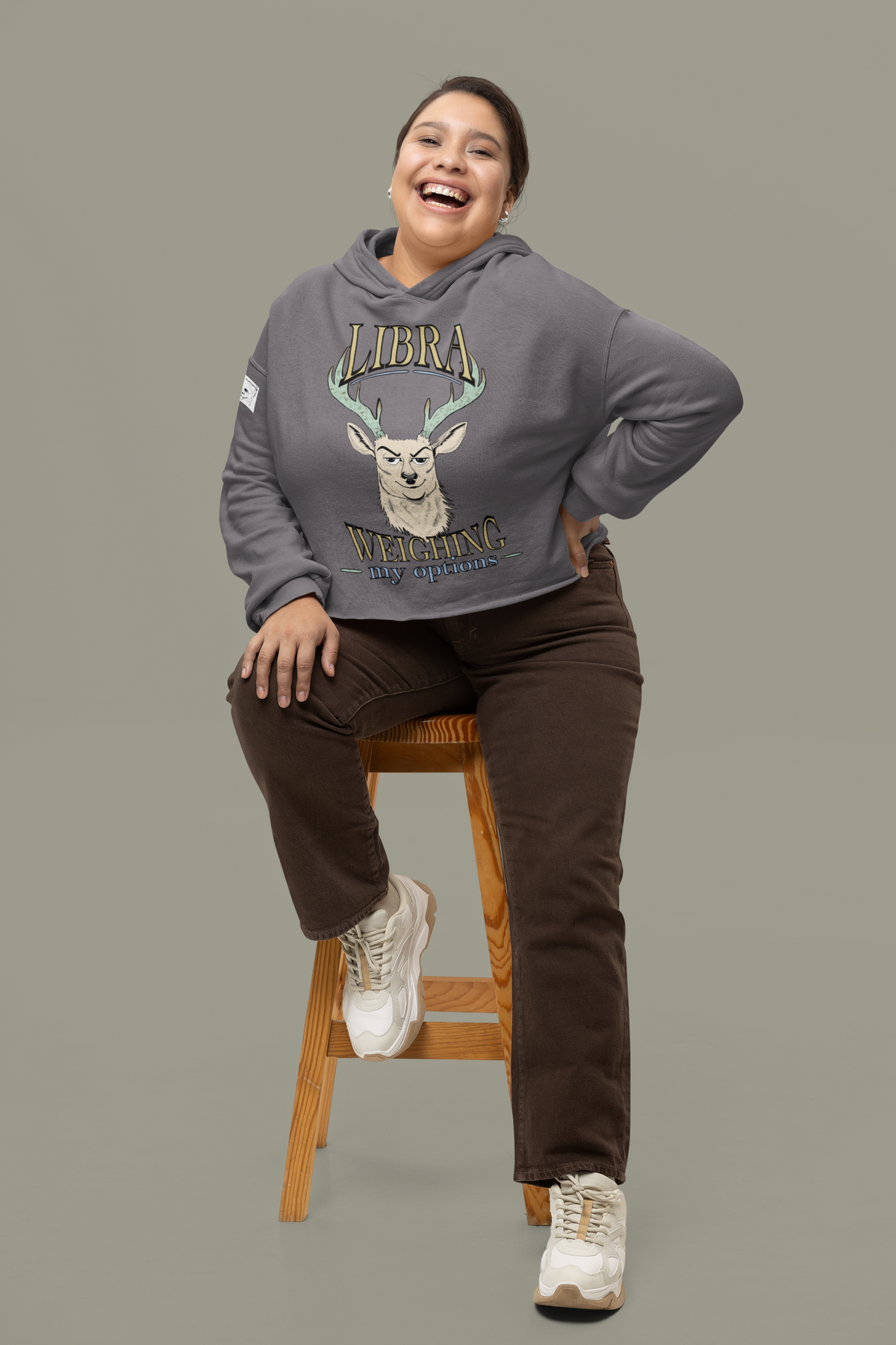 Woman sitting on a wooden stool in a storm grey cropped Libra Weighing My Options hoodie, cream stag with mint antlers and gold lettering.