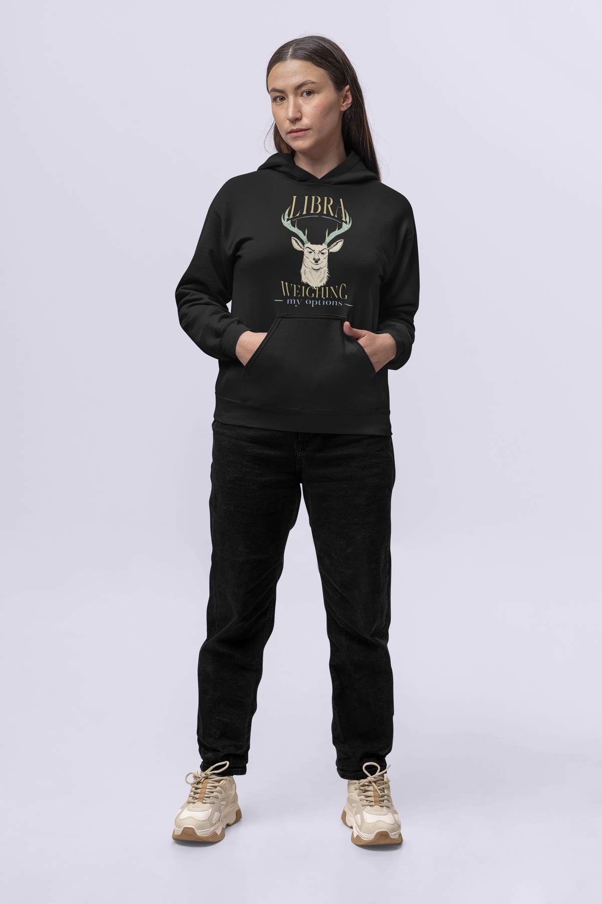 Woman standing with hands in the pockets of a black Libra Weighing My Options hoodie, cream stag, mint antlers, and gold lettering.