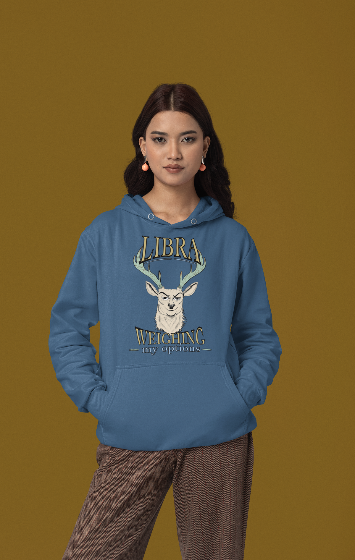 Woman in a studio wearing an indigo blue Libra Weighing My Options hoodie, cream stag with mint antlers and gold lettering.