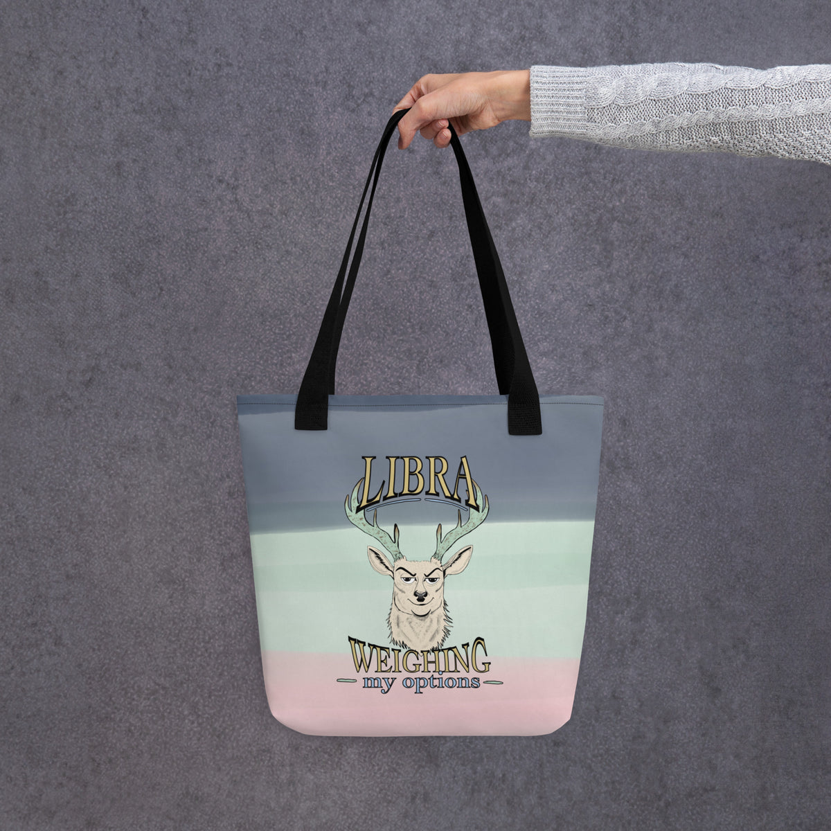 Back view of the Libra Weighing My Options all-over print tote held by a hand, cream stag with mint antlers and gold lettering on a mint-to-pink watercolor wash with black straps.
