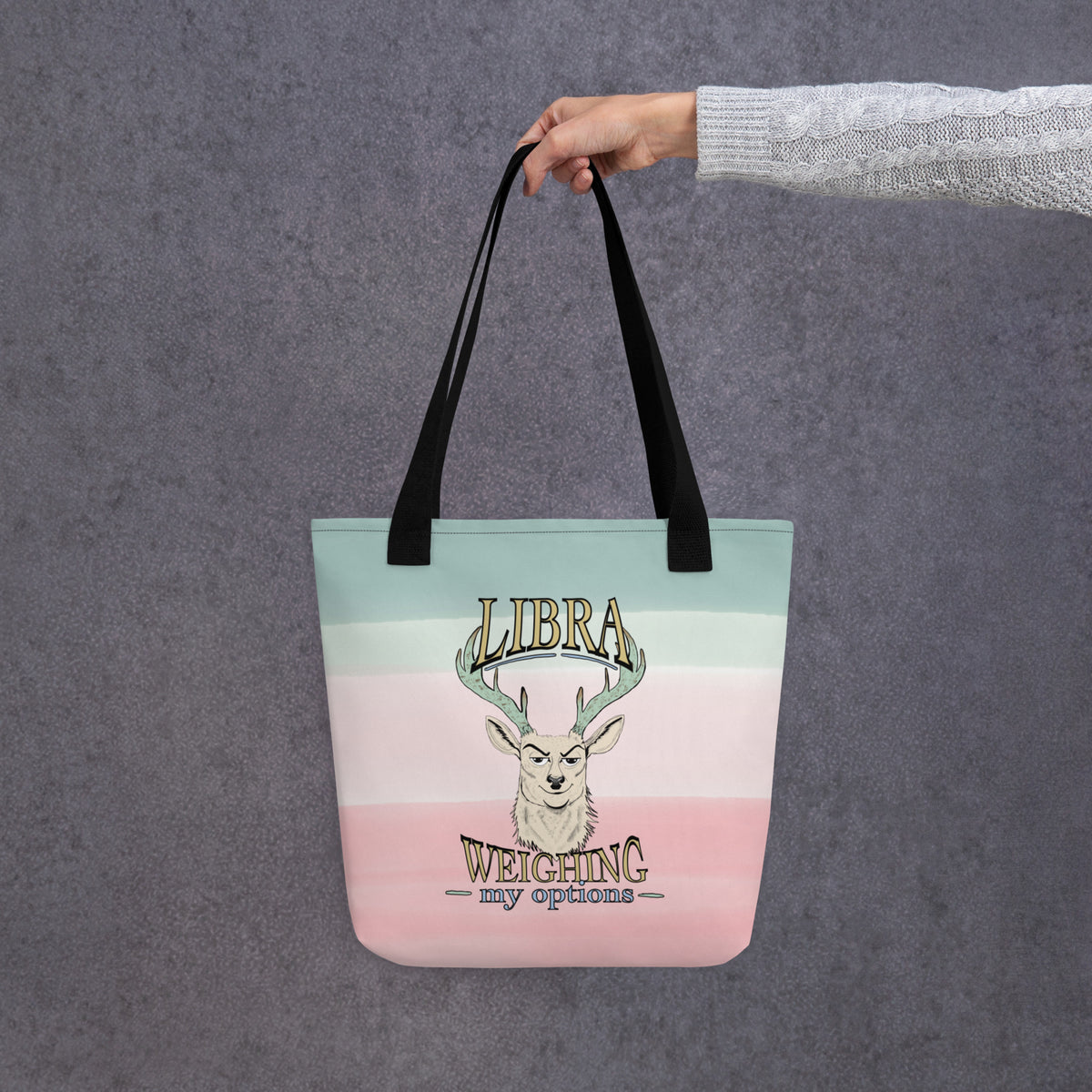 Front view of the Libra Weighing My Options tote held by a hand, cream stag with mint antlers, gold LIBRA and WEIGHING my options lettering on a mint-to-pink watercolor wash.