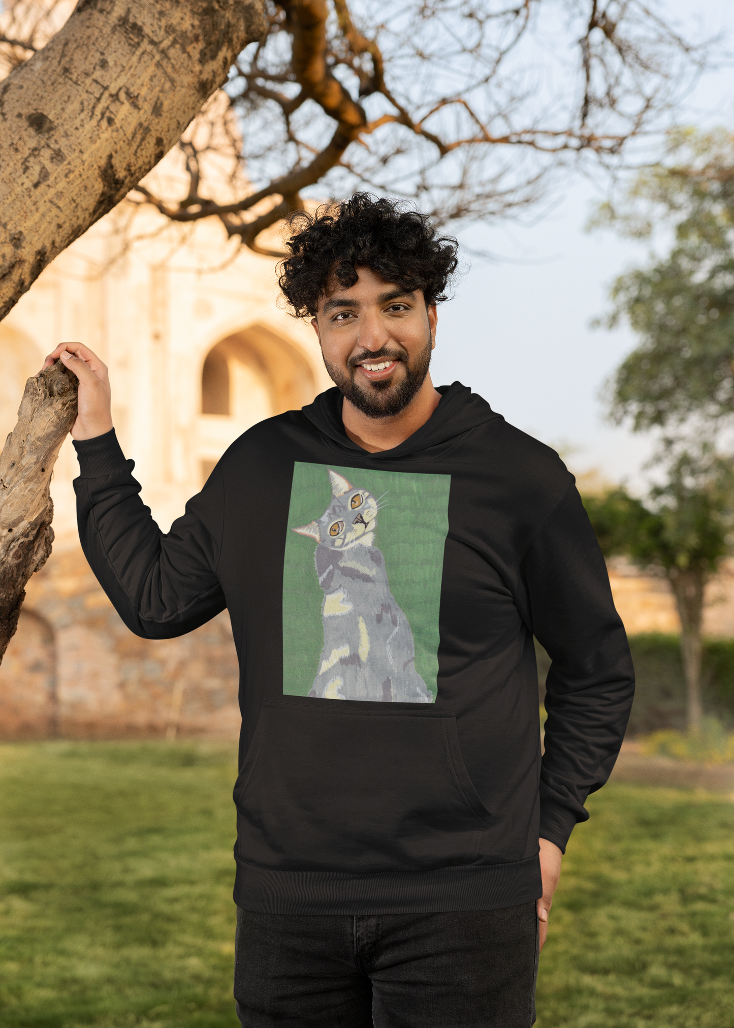 Man with curly hair standing outdoors next to a tree wearing a black hoodie with Mr. Grey cat portrait