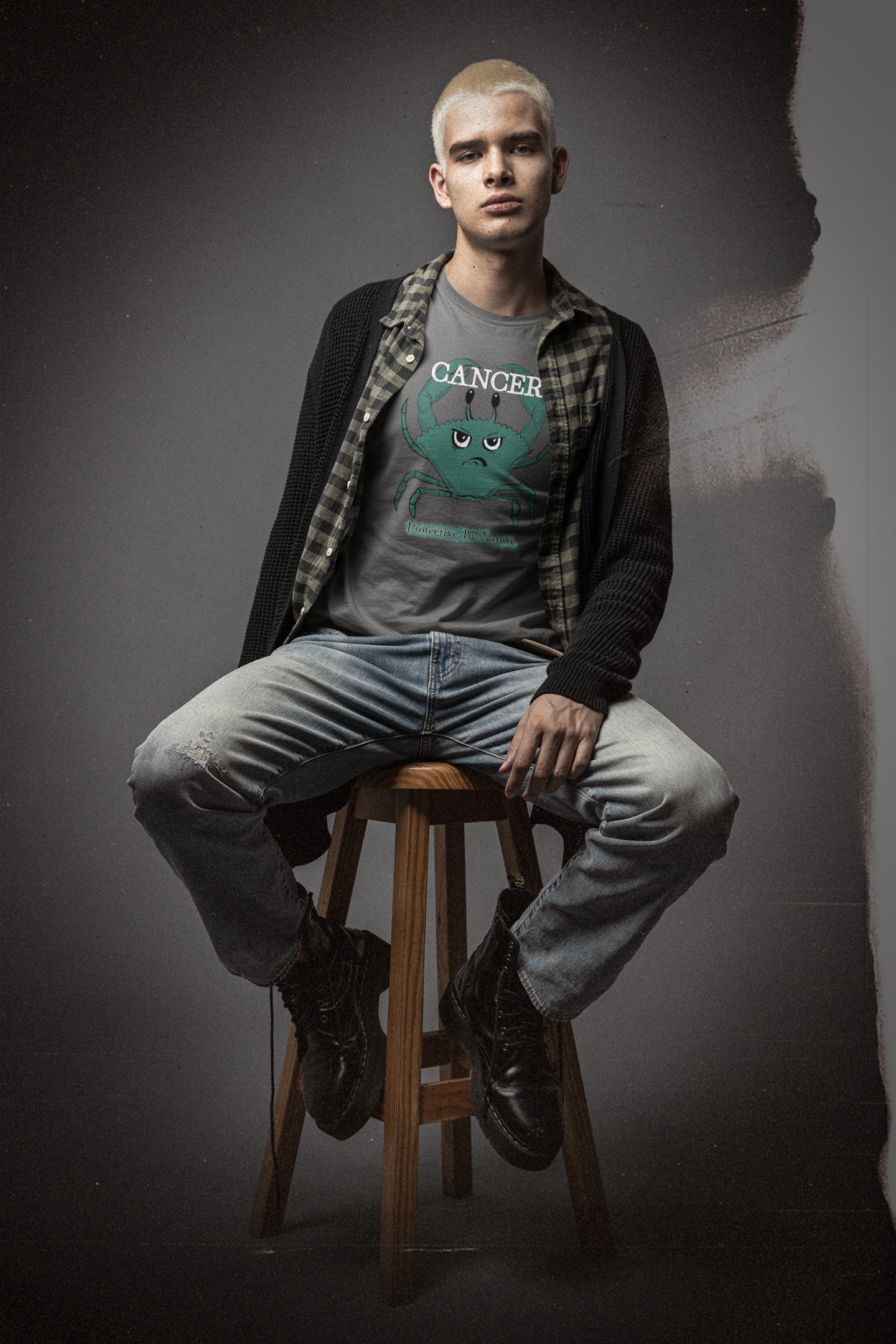 Young man with platinum blonde hair wearing an asphalt grey Cancer "Protective By Nature" t-shirt sitting on a wooden stool in a moody studio