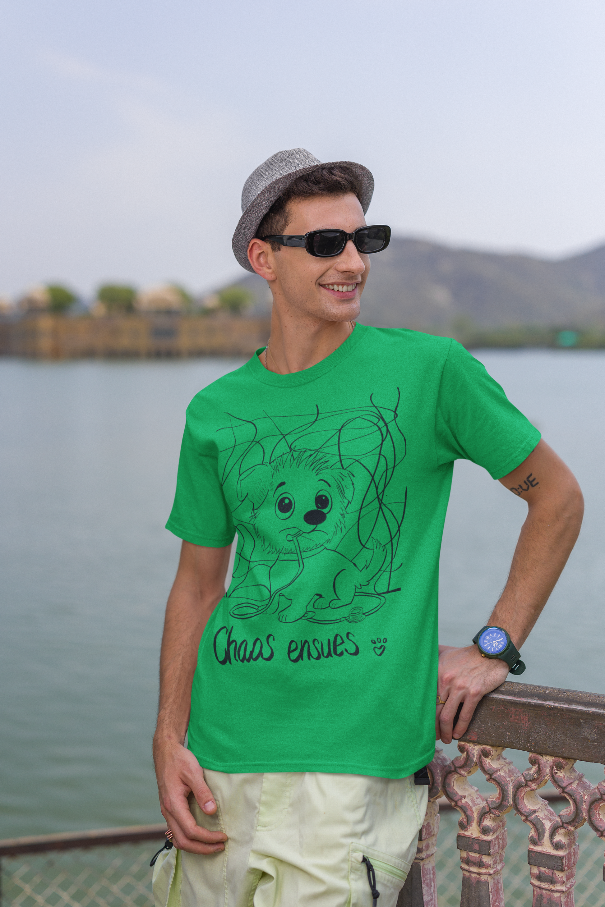 Smiling man wearing green Chaos Ensues t-shirt standing by water with sunglasses and hat