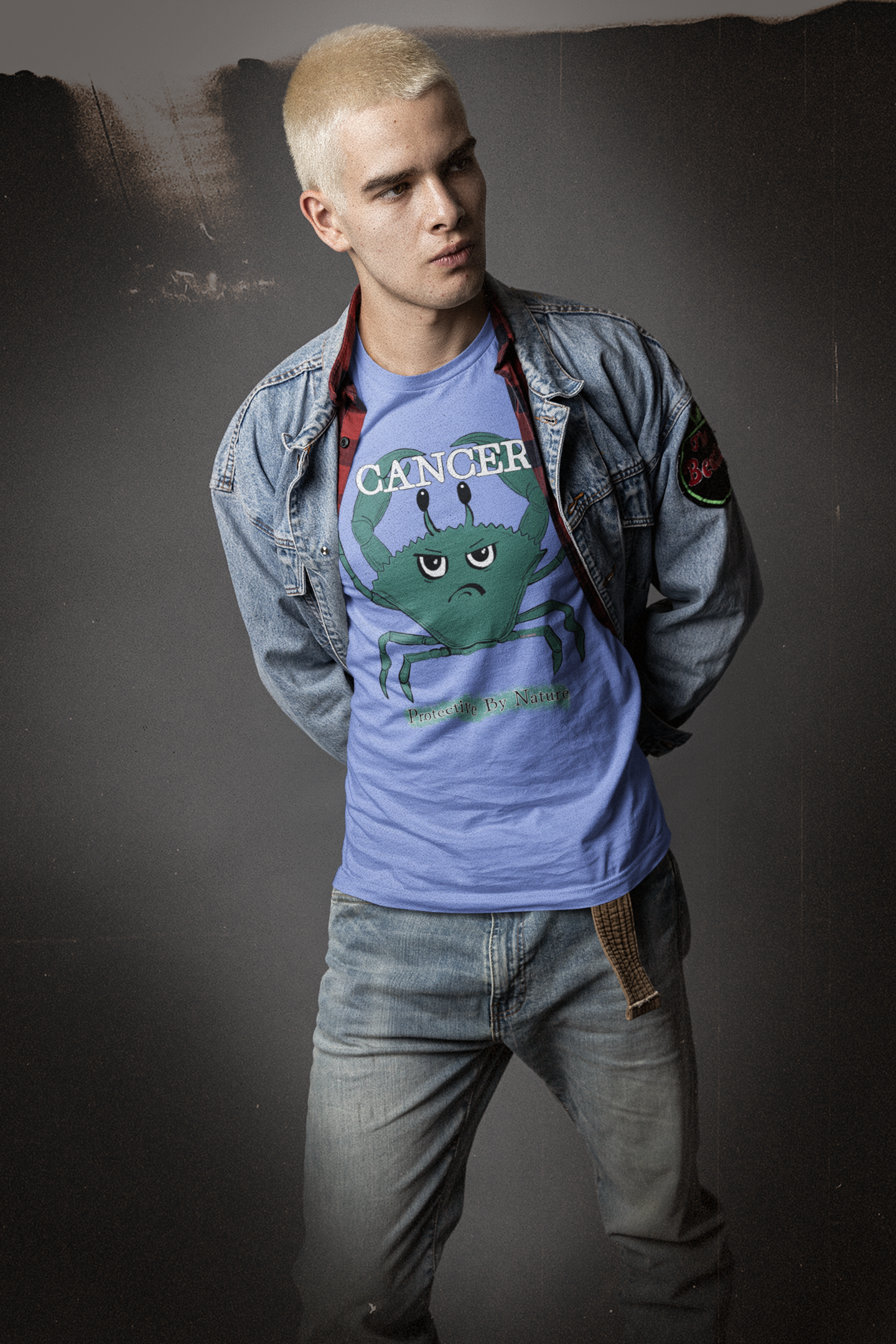 Young man with platinum blonde hair wearing a heather columbia blue Cancer "Protective By Nature" t-shirt under a denim jacket, posing against a dark background