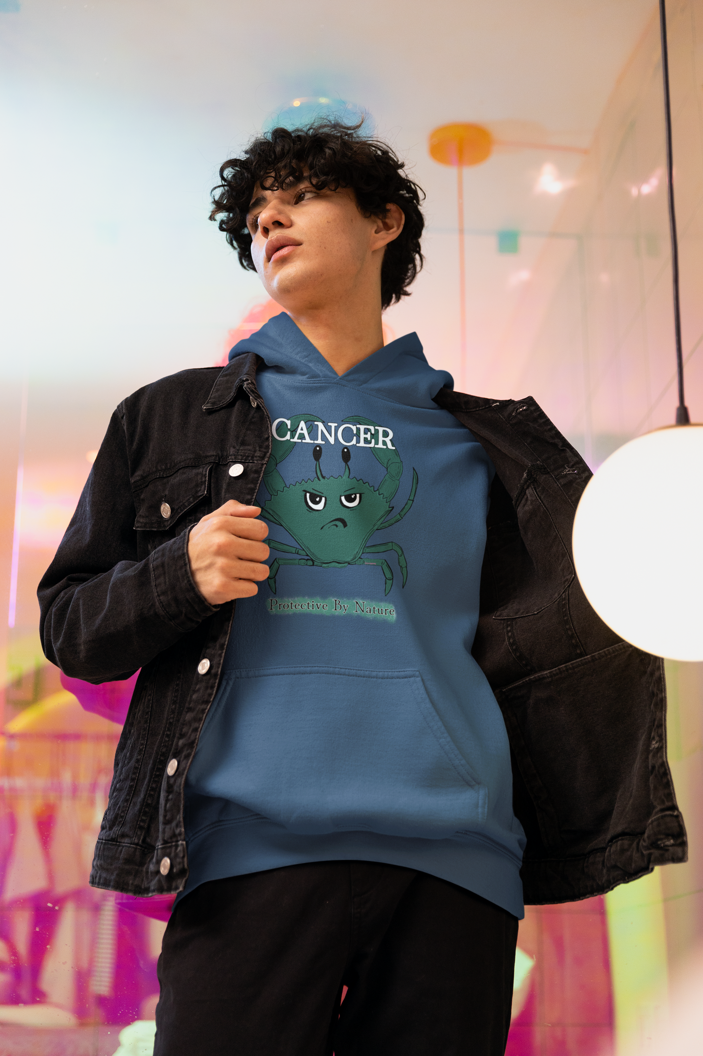Young man with curly hair wearing an indigo blue Cancer "Protective By Nature" hoodie under a black denim jacket, standing in a vibrant studio