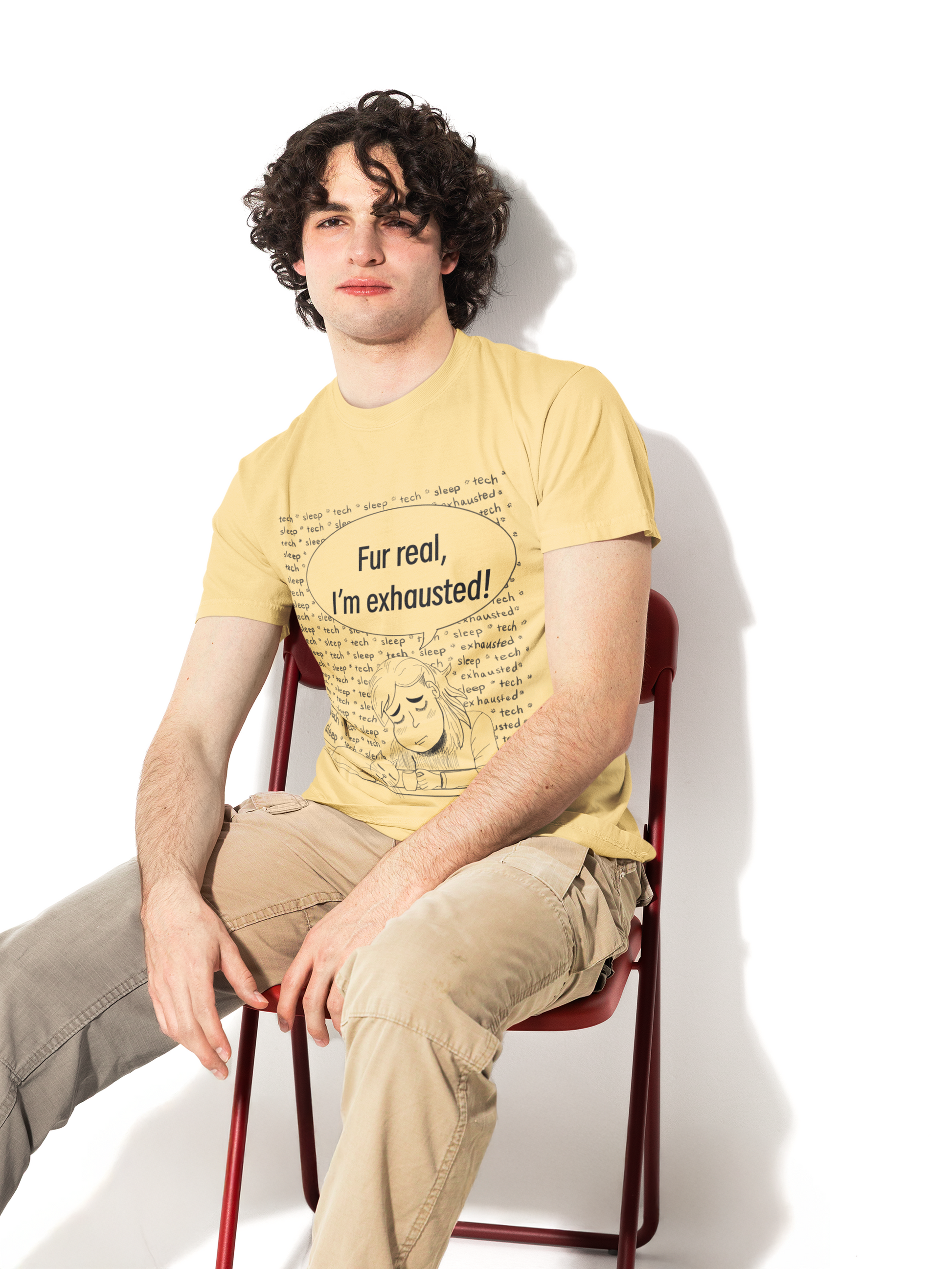 Man with curly dark hair wearing a butter yellow Fur Real, I'm Exhausted t-shirt sitting on a red chair