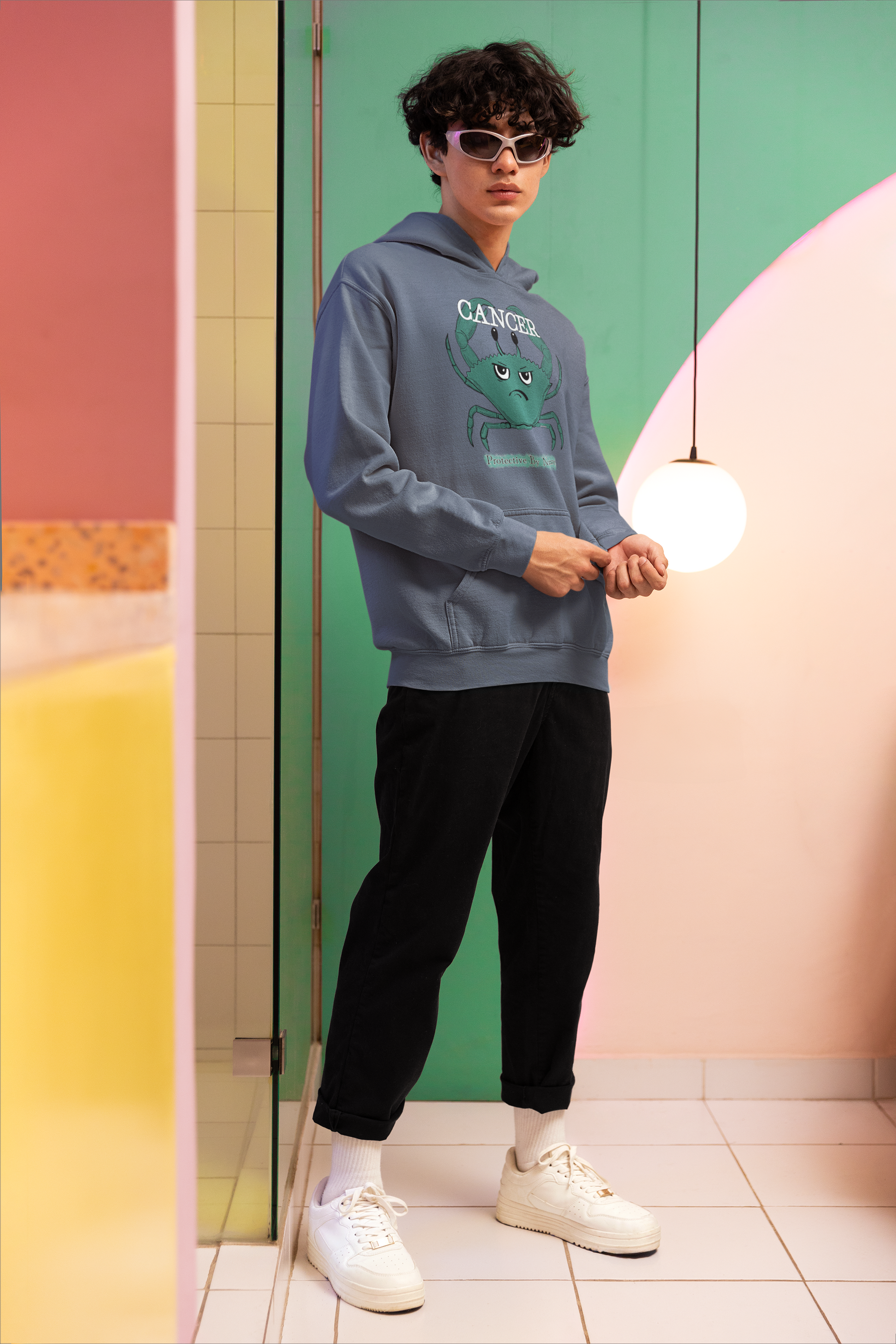 Young man with curly hair wearing a heather sport dark navy Cancer "Protective By Nature" hoodie, posing in a colorful modern setting