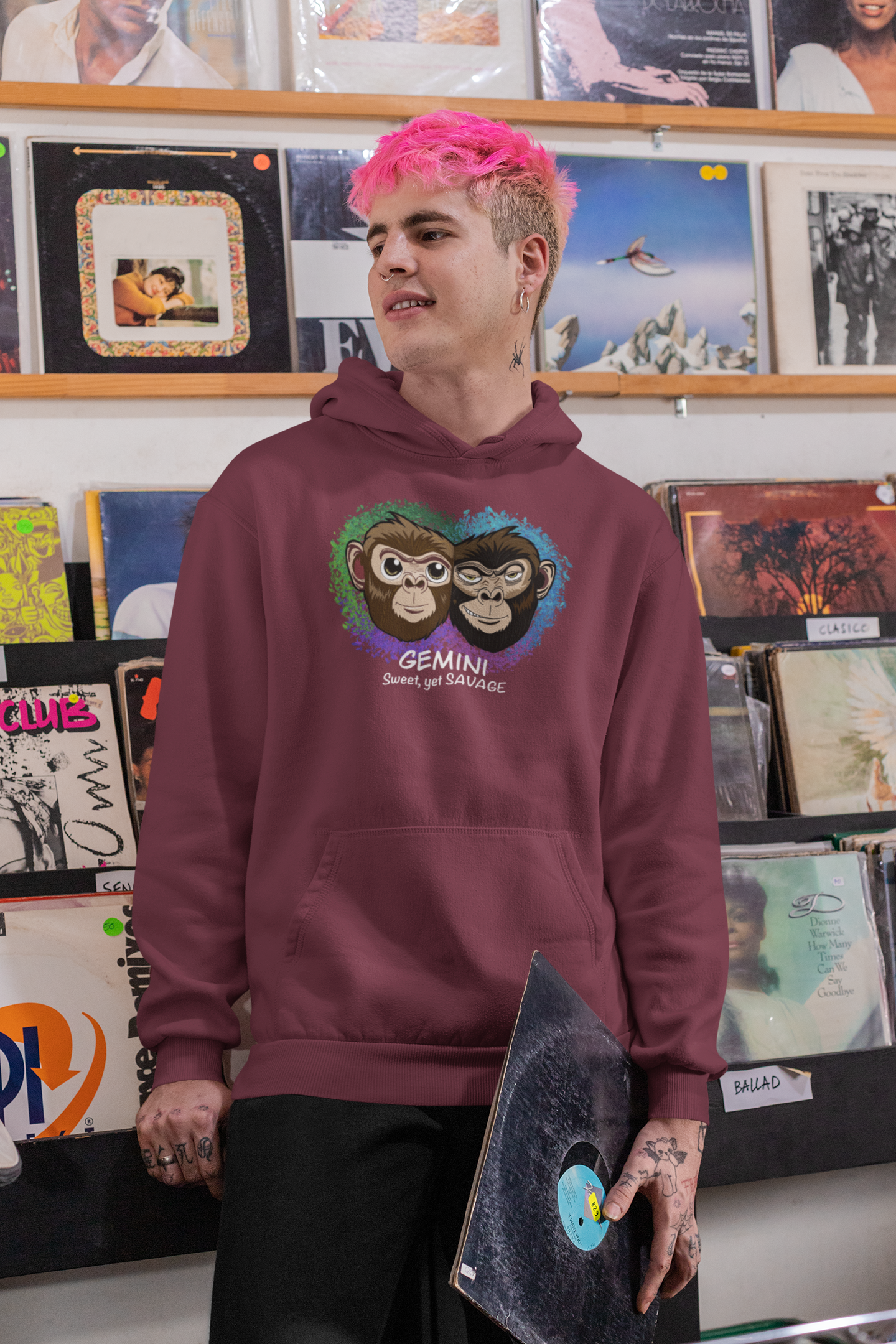 Man with bright pink hair wearing a maroon Gemini "Sweet, yet SAVAGE" hoodie posing in a record store with vinyl records behind him