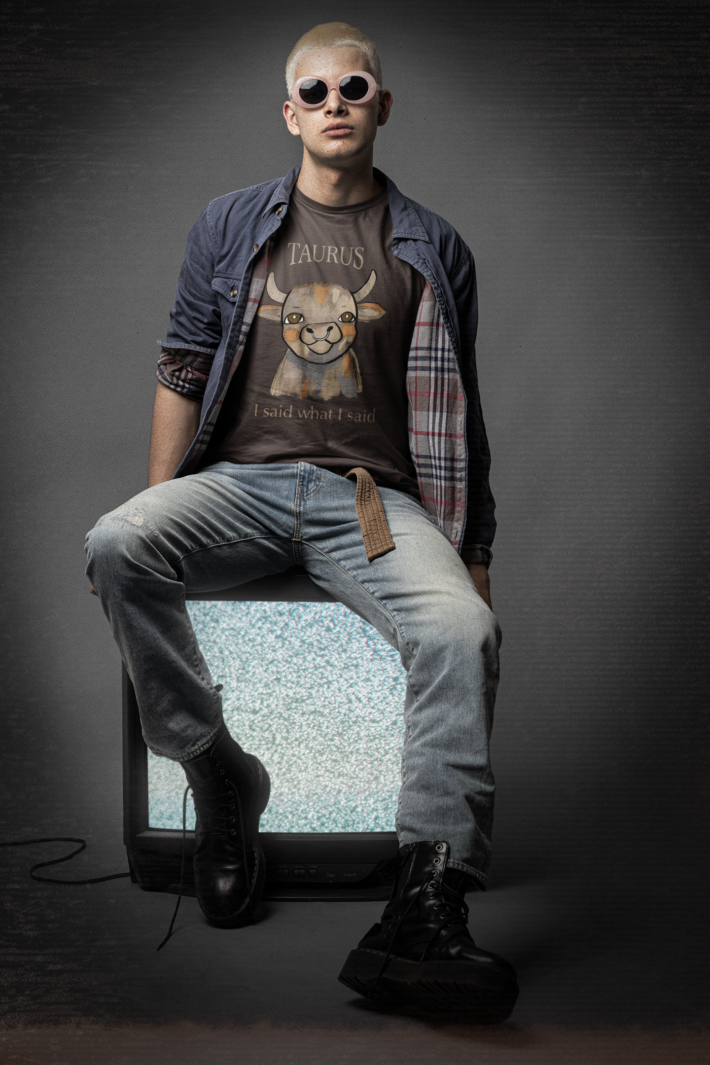 Man with bleached hair and pink sunglasses sitting on a TV wearing a brown Taurus Bull t-shirt