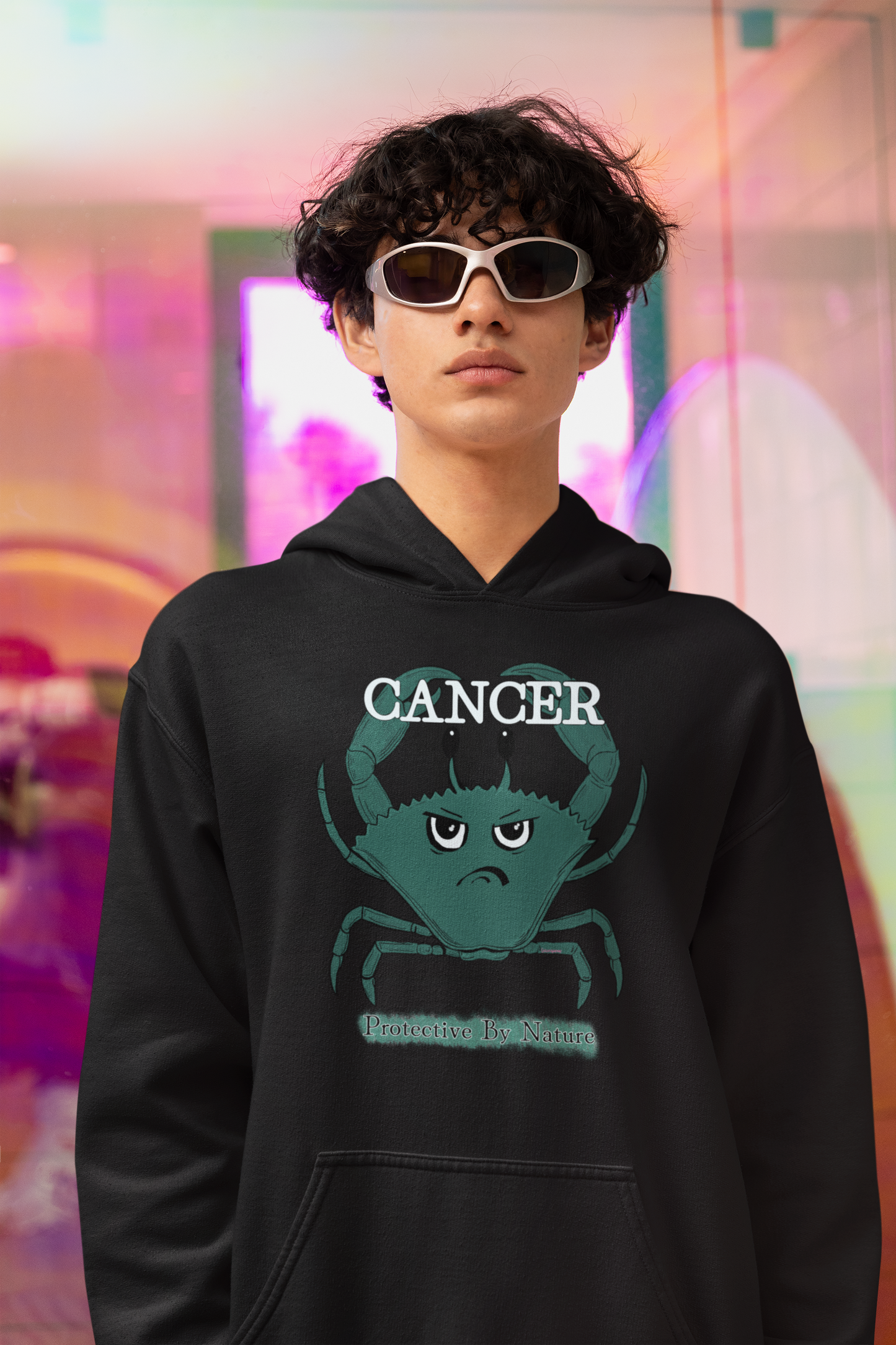 Man with curly hair and sunglasses wearing a black Cancer "Protective By Nature" hoodie under a black denim jacket, posing with a serious expression